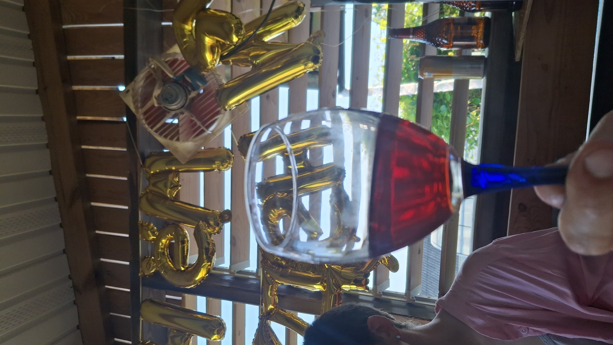 This media file shows a close-up of a person's hand holding a wine glass filled with red liquid. The wine glass has a blue stem. In the background, there are gold foil balloons shaped like numbers, and a small fan with red and white blades. Behind the balloons, there are wooden structures and sunlight filtering through slats, suggesting an outdoor or semi-outdoor setting. There are also two bottles visible on a shelf in the background, one amber glass and one clear glass. The scene appears to be a celebration or a casual gathering, possibly outdoors given the natural light. The lighting suggests it is daytime. There is no visible text in the image. The overall impression is festive and relaxed.