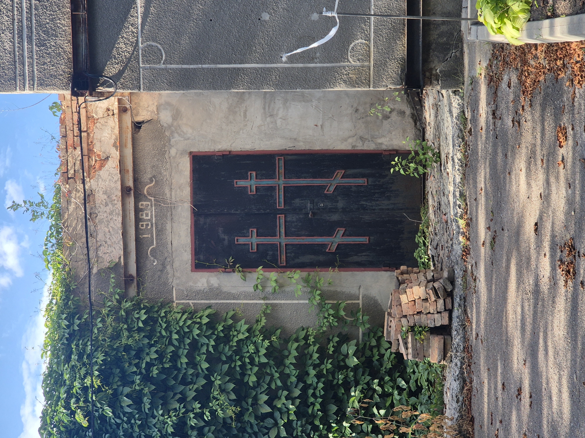 The image shows a weathered, dark double door with red trim, adorned with two ornate crosses featuring a distinctive lower crossbar, possibly representing Eastern Orthodox crosses. The door is set into a grey, textured concrete facade, which is partially overgrown with lush green ivy on the left side. Above the door, a brick archway shows signs of age and decay, with exposed red bricks and crumbling mortar. The number "1989" is embossed in the concrete above the archway. To the left of the door, a pile of used red bricks is stacked on a cracked concrete surface. The ground in front of the structure is asphalt, covered with fallen leaves. To the right of the door, a vertical white ribbon or tape is attached to the grey facade with a thin wire. The overall scene suggests an abandoned or neglected structure within a cemetery or an old building in Sozonivka, Ukraine, under a bright, partly cloudy sky.