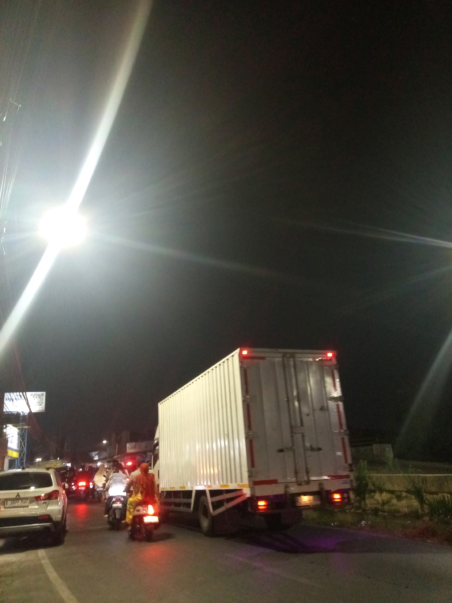 This image depicts a nighttime scene of traffic congestion in the city of Bekasi, Indonesia. In the foreground, a white car is stationary. Behind it, several motorcycles are also stopped. One motorcycle has two occupants, one of whom is wearing a yellow shirt and patterned trousers, and the other is wearing a white shirt. Further down the road, a large white truck with a long trailer is visible, also appearing to be in traffic. The sky is dark, with a bright light source illuminating the left side of the frame, possibly a streetlamp or headlights. There are also streetlights and building lights visible in the background. The road surface is asphalt. The overall impression is one of a busy street at night, with vehicles and people navigating through traffic.