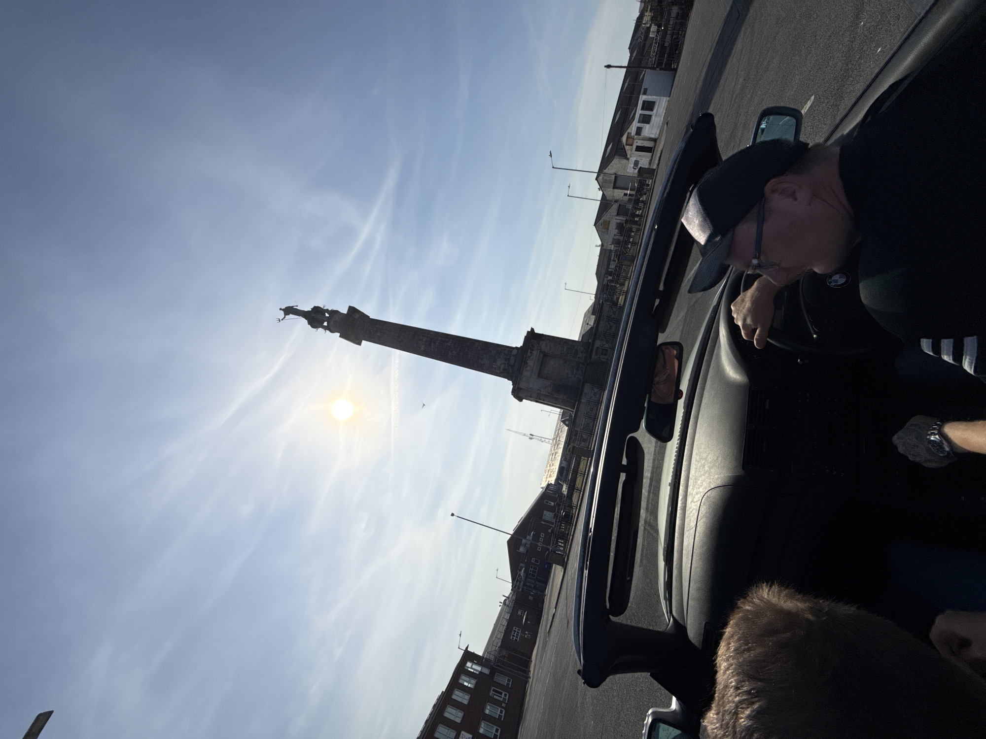 The image shows a partially-framed view of a man in a car, with Nelson's Monument in Great Yarmouth, United Kingdom, visible through the windshield. The man is wearing a baseball cap and glasses, and appears to be in the driver's seat. The sun is shining behind the monument, a tall, fluted Doric column topped with a statue of Britannia. Buildings and streetlights can be seen in the background beyond the monument, indicating an urban setting. The image is taken from inside the car. The car's dashboard, steering wheel, and mirror are partially visible. The weather appears to be sunny with a mostly clear sky.