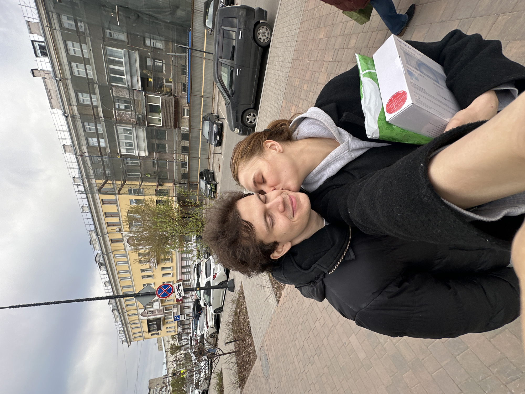 Two people are standing on a brick-paved area next to a street with parked cars in Krasnoyarsk, Russia. A woman is kissing a man on the cheek. He holds a white box and a green bag. Buildings are visible in the background. Construction work with scaffolding is on one building. Street signs are nearby.