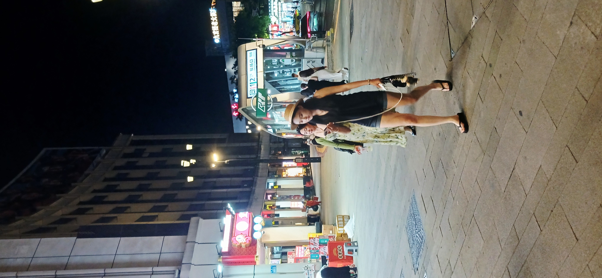 The image shows two women standing on a sidewalk in Yuzhong District, China at night. One woman is wearing a dark top, light-colored shorts, and a straw hat, while the other is wearing a light-colored floral dress and a green cardigan. The woman in the shorts is holding what appears to be a toy gun with a white wire attached. In the background, there are stores and a building with lights. A bus stop with Mandarin characters is visible. The sidewalk is made of light-colored stones.