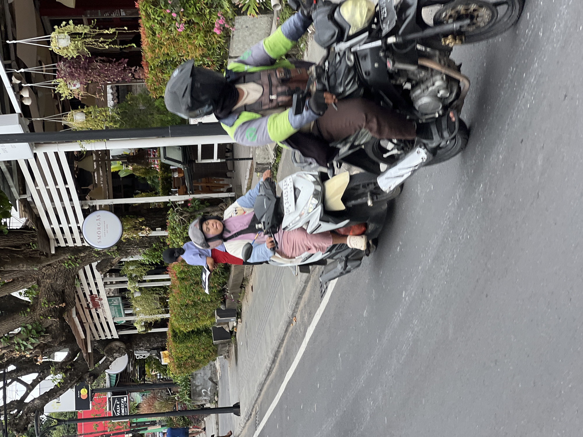 The image depicts a street scene in Denpasar, Indonesia, with two people riding motorcycles. The foreground shows the rear of a black motorcycle with a rider wearing a helmet, a green and gray jacket, brown pants, and black gloves. The motorcycle has a license plate visible. To the left, a white motorcycle is seen with a rider wearing a helmet, a pink and white jacket, and pink pants. This motorcycle also has a visible license plate. In the background, there are buildings with white striped awnings, trees, and lush green bushes. A sign for "SMORGAS" is visible on one of the buildings, and another sign for "YULIA 2 HOMESTAY" is also present. The street is paved, with a sidewalk to the left. The overall atmosphere suggests a typical urban environment in Bali.
