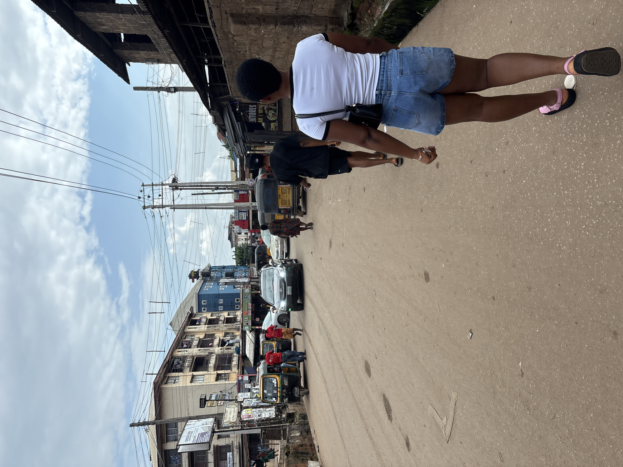 In the city of Onitsha, Nigeria, a street scene unfolds under a cloudy sky. The foreground features a woman walking away from the camera, her back to the viewer. She wears a white t-shirt with black trim, denim shorts, and black sandals with pink straps. A black purse hangs from her shoulder. Behind her, a man in a black t-shirt and shorts walks away, wearing a black cap. Further down the street, a yellow auto-rickshaw is parked, with two men in red t-shirts standing beside it. A silver car is moving towards the camera, and other vehicles and buildings line the street. The road appears to be made of dirt and gravel. The environment suggests a bustling urban area with typical street life.
