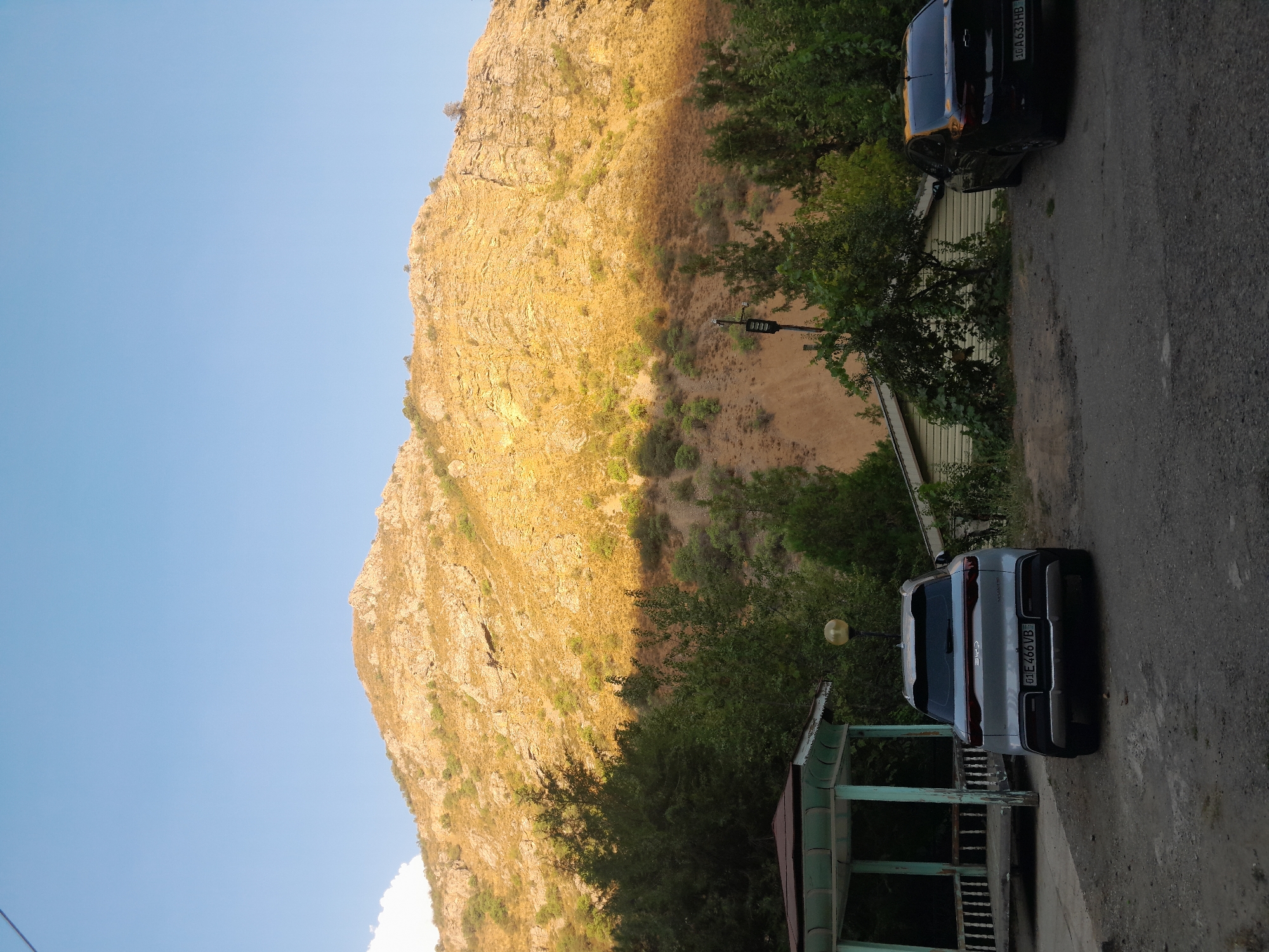The image depicts an outdoor scene in an area with a mountainous backdrop. In the foreground, there are two cars parked on a dirt and gravel surface. The car on the left is a light-colored SUV, and the car on the right is a black sedan. To the left of the SUV, there is a light-colored gazebo with a green roof and decorative white railings. Behind the cars and gazebo, there is a building with a light-colored, horizontal siding. A tall pole with a light fixture is visible between the cars. The middle ground is filled with lush green trees and foliage, with some taller evergreen trees. The background is dominated by a large, rocky mountain slope, illuminated by warm sunlight, casting a distinct shadow across a portion of the slope and the trees. The sky above is clear and blue, with a few faint clouds on the far left. The overall impression is of a natural landscape with some man-made structures and vehicles.