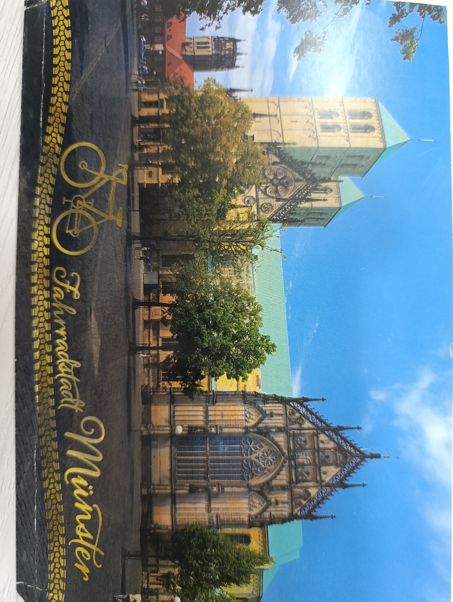 The image shows a postcard featuring a picturesque view of a historic European city, likely a cathedral or a similar large, ornate religious building with prominent towers and a green roof. The architectural style suggests Gothic influences. In the foreground, to the left, a golden bicycle silhouette is depicted against a dark background with a tire track pattern. Vertical golden text reads "Fahrradstadt Münster," which translates to "Bicycle City Münster." The scene is set against a clear blue sky with some wispy clouds, indicating a sunny day. Trees with green foliage are scattered around the buildings, suggesting an urban park or street setting.
The prompt states to include the location in the city of Termez, Uzbekistan. However, the visual information in the image (architectural style, text, and overall aesthetic) does not align with any known landmarks or characteristics of Termez, Uzbekistan. Termez is known for its ancient Buddhist and Islamic archaeological sites, which have a distinct architectural appearance different from what is shown in the postcard. The text "Fahrradstadt Münster" explicitly identifies the location as Münster, Germany, a city renowned for its cycling culture. Therefore, it is impossible to provide details about Termez, Uzbekistan based on this image.