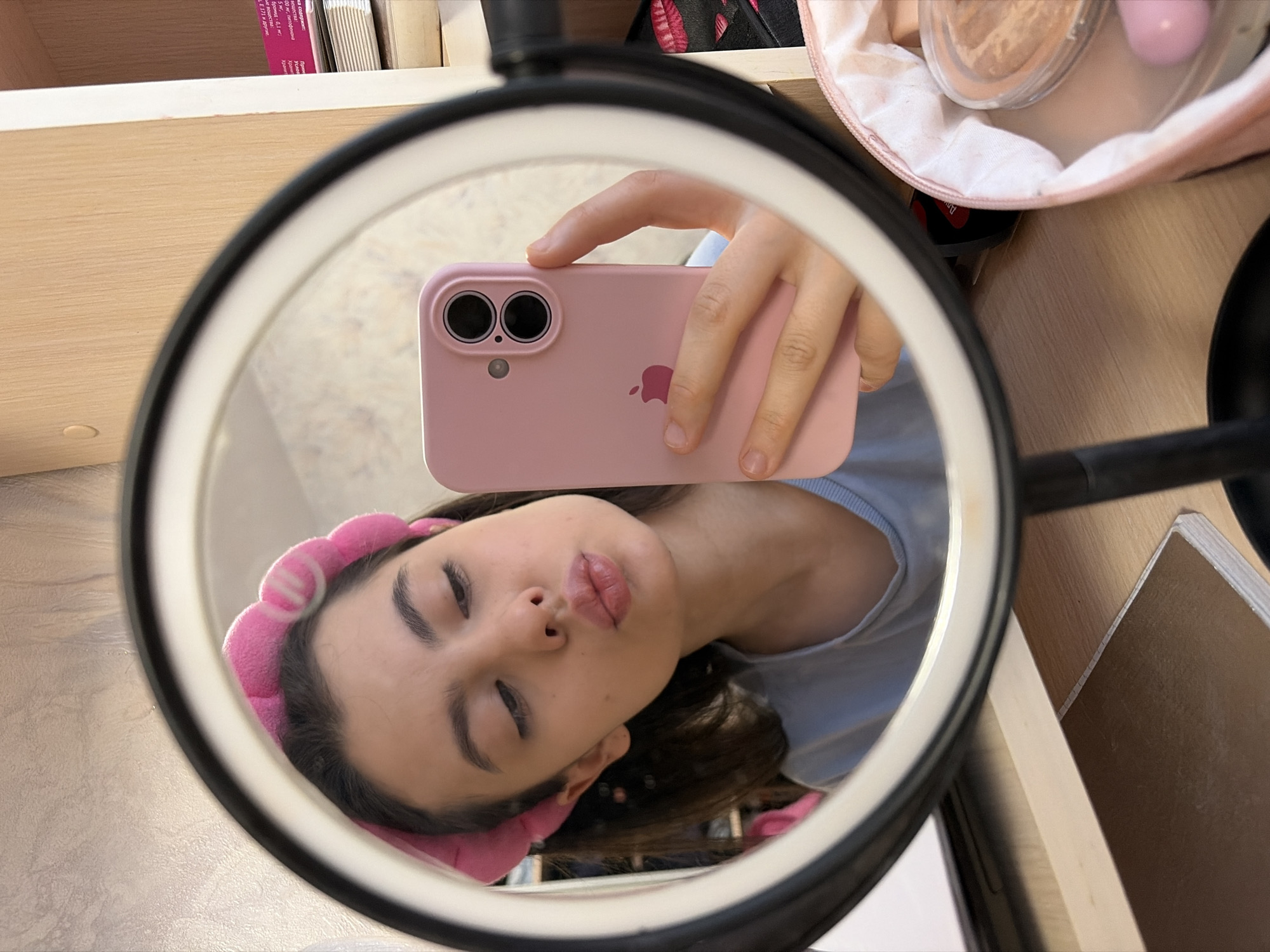 A woman wearing a pink headband and a light blue shirt is reflected in a ring light-mounted mirror, holding a pink iPhone in front of her face. The mirror is set on a light-colored wooden surface where a makeup bag, cosmetics, and books are also visible. The city of Chaikovsky, Russia, is not discernible from this image.
