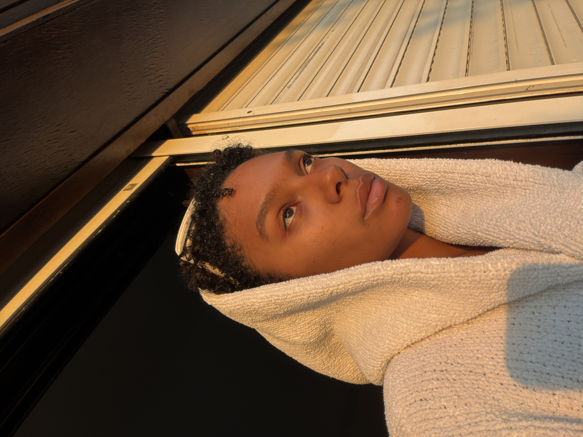 A young person, whose face is partially obscured by a white hooded robe, gazes outward from a window frame. The subject's dark, curly hair is visible beneath the hood, and their skin is illuminated by the warm glow of golden hour sunlight. The background includes the dark interior of a room and the exterior of a building with closed shutters. The overall impression is one of contemplation or observation, possibly from an apartment or residential building in Champs-sur-Marne, France.