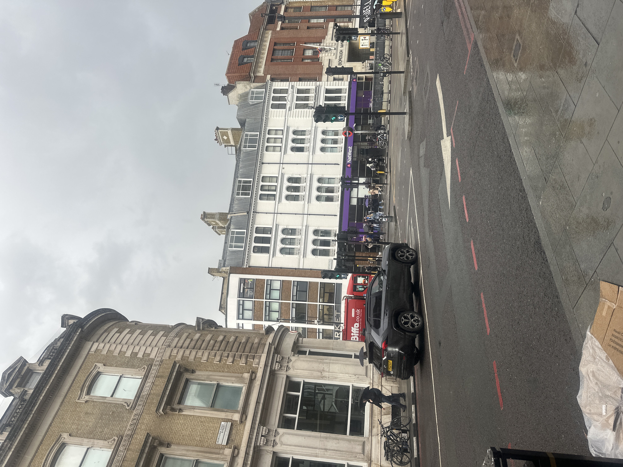 The image depicts a street scene in London, United Kingdom, likely near Borough Market. A grey SUV is parked on the right side of the road, while a red Biffa waste management truck is visible further down the street. Pedestrians are walking on the sidewalk, some with umbrellas, indicating overcast or rainy weather. The buildings are a mix of architectural styles, with some featuring ornate detailing and others being more modern. Traffic lights are visible, suggesting an intersection. The road surface appears wet, reflecting the streetlights. The sky is overcast and grey.