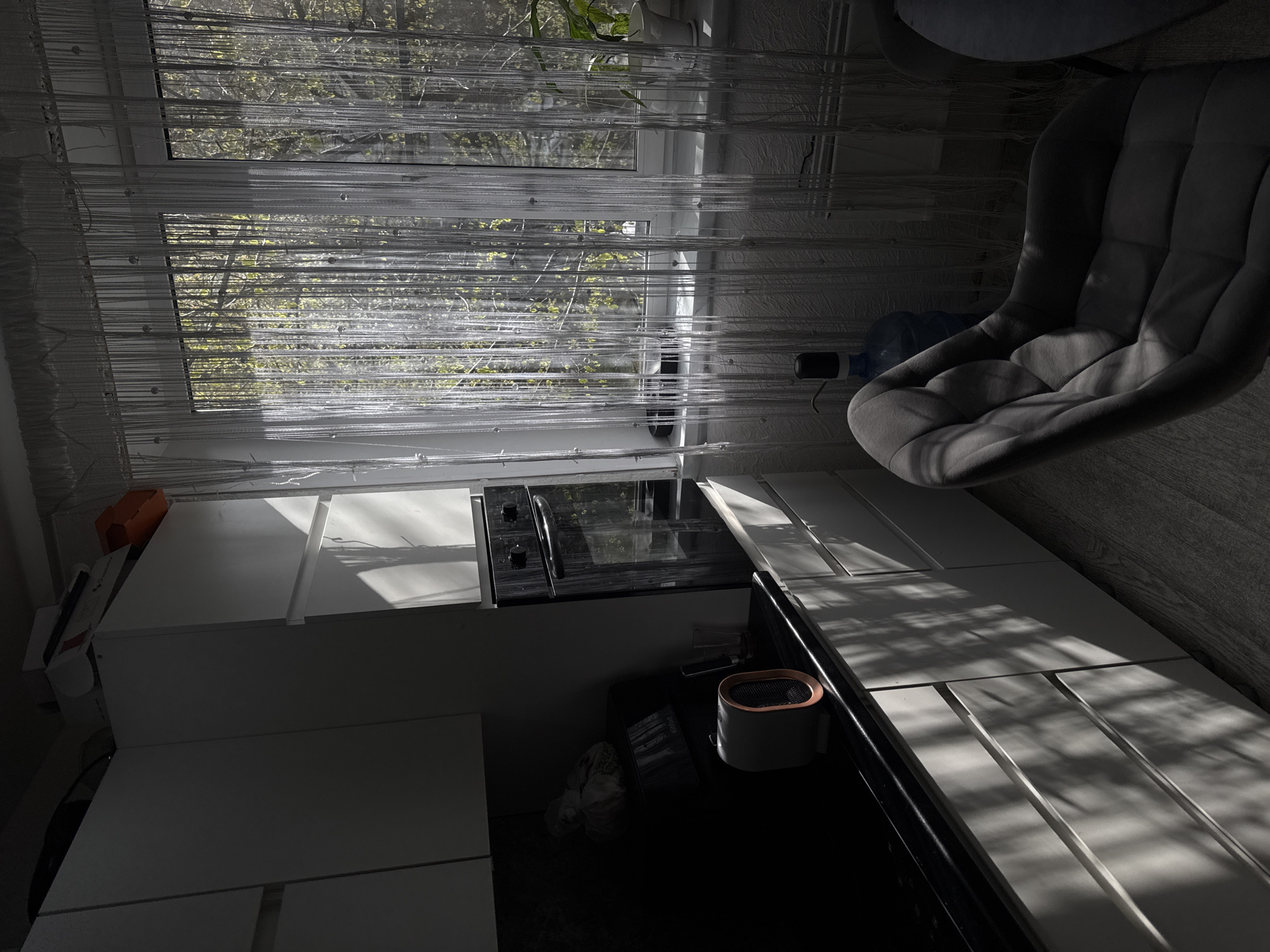 The image shows an interior view of a room, possibly a kitchen or dining area. The dominant features include white kitchen cabinets, an oven, and a gray cushioned chair. A window with string curtains is visible in the background, with a view of trees and greenery. A water bottle is placed near the chair. The lighting is strong, with sunlight casting shadows across the scene. A small kitchen appliance is positioned on the black countertop.