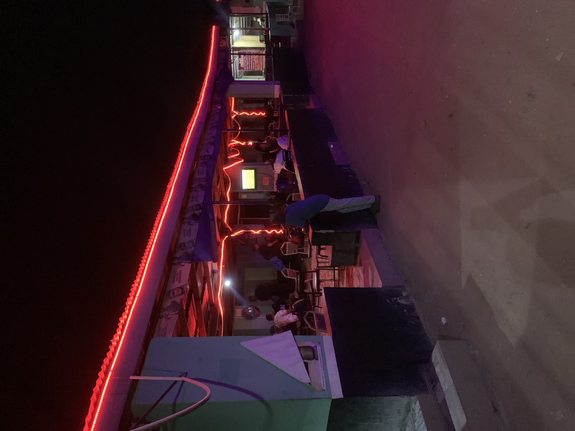 The scene depicts an outdoor bar or eatery at night, possibly in Kabong, Nigeria. The building's roofline is illuminated with a continuous red LED strip. Underneath, people are seated at tables, suggesting a social gathering. Some individuals appear to be interacting. A person is bending over near the center of the frame. A television screen can be seen. The surrounding ground appears to be unpaved or a rough road surface. The image quality is low, suggesting it might have been taken with a mobile device. The overall lighting is dim, with the red LEDs providing the primary source of illumination.