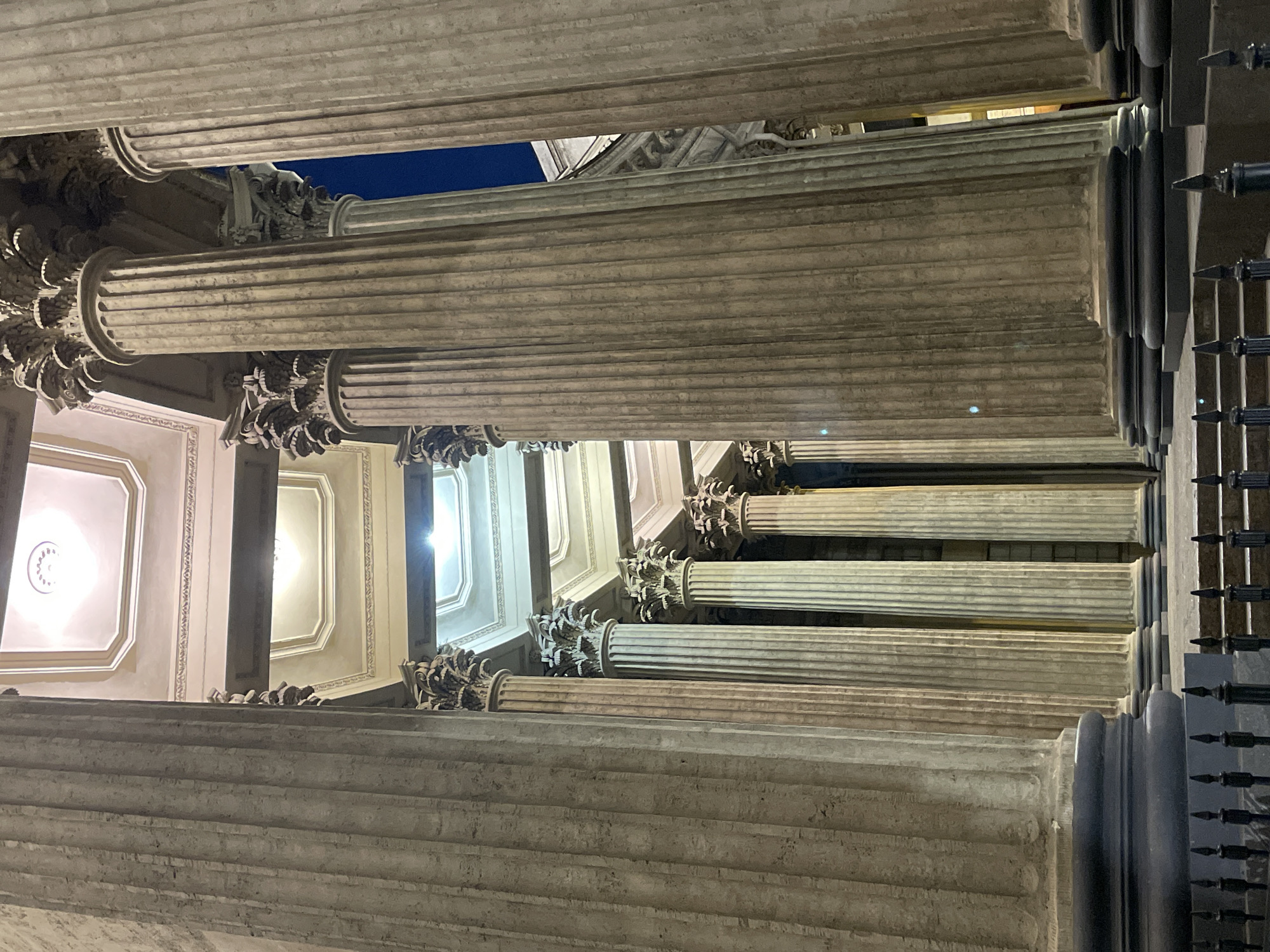 The image shows a low-angle view looking up at a series of Corinthian columns. The columns are made of light-colored stone. Ornate carvings can be seen at the top of the columns. Behind the columns, sections of the ceiling are visible, decorated with geometric patterns and central light fixtures. Glimpses of dark blue sky appear between the columns and the top of the building. To the right edge of the image is a black fence. The location is likely a building in Saint Petersburg, Russia, given the architectural style. There are no people visible.