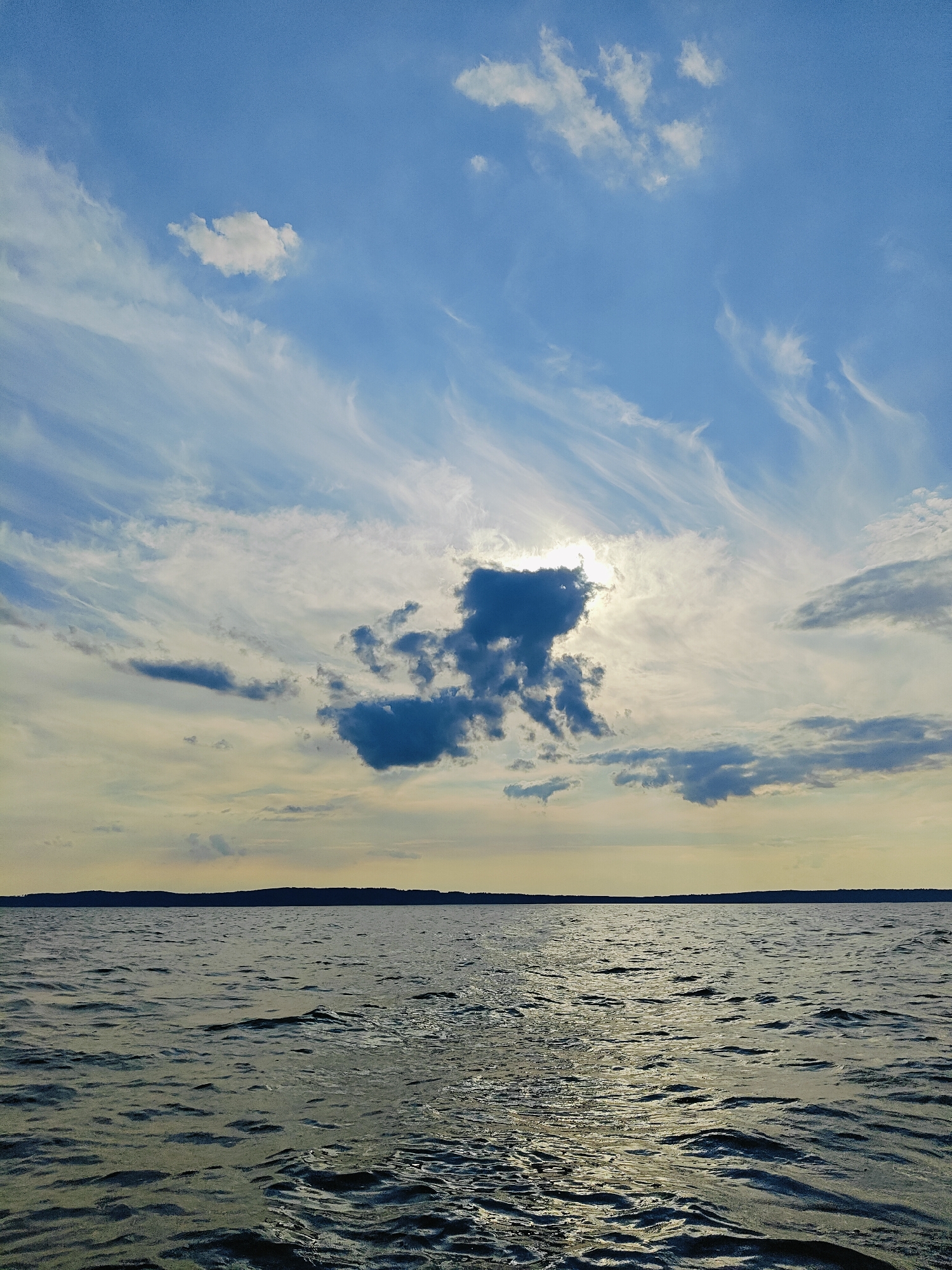 The image shows a wide expanse of water, likely a lake or river, with a dark, silhouetted tree line in the distance. The sky above is a mix of blue and scattered clouds, with the sun partially obscured by a dark, cumulus cloud in the center. The water has a choppy surface, reflecting the sky and sunlight. The scene appears to be a natural landscape, possibly a waterfront area in or near Chaykovsky, Russia, given its typical geography. There are no people or specific objects visible that would indicate a particular activity or event. The overall impression is one of natural beauty and a calm, open environment.