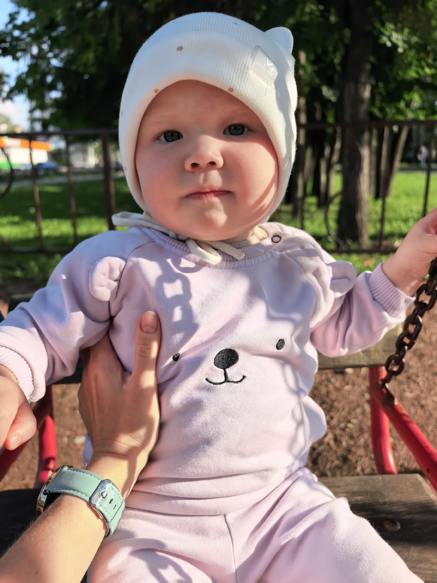 A young child, appearing to be an infant or toddler, is seated on a wooden swing with red metal supports, outdoors in what appears to be a park or playground in Sterlitamak, Russia. The child, with fair skin and blue eyes, wears a white knitted beanie adorned with small light brown polka dots and two rounded ear-like protrusions, featuring a small white "Smile" label on the left side. The child is dressed in a light lavender or pale pink long-sleeved hooded outfit, which includes bear ears on the shoulders and a black embroidered bear face (nose and mouth) on the chest. An adult's hand, with fair skin, is visible supporting the child's left side/upper arm. The adult wears a watch with a light teal or mint green leather strap and a silver buckle on their left wrist. The child looks directly forward, towards the camera. Strong sunlight illuminates the scene, casting distinct shadows, such as on the child's outfit and the adult's hand. In the blurred background, green grass, dark tree trunks, and foliage are visible behind a brown metal fence or railing. Parts of other playground equipment or distant structures are faintly discernible in the upper left, beneath a light blue sky. A thick metal chain from the swing set is visible on the right side of the frame. The ground beneath the swing appears to be dark brown, possibly wood chips or dirt.