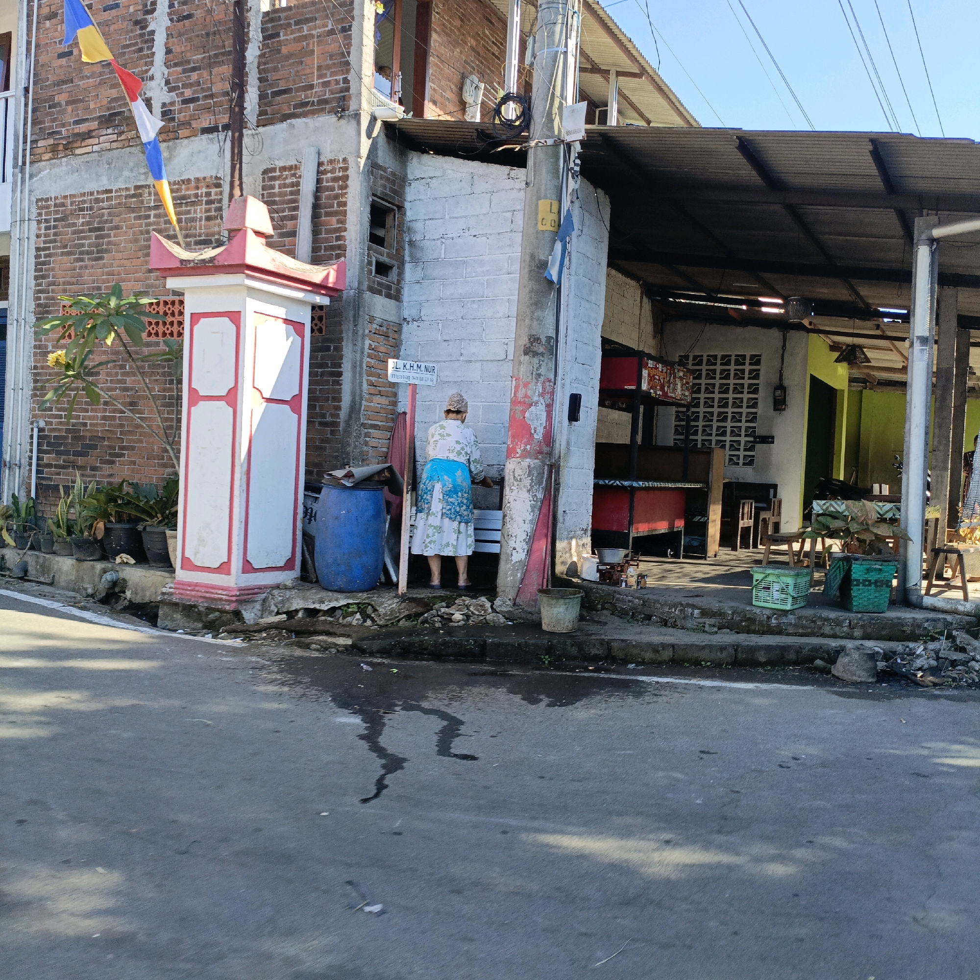 An outdoor, daytime scene in the city of Jombor, Indonesia, depicts a street in front of a small building with a covered outdoor area. A woman, wearing a patterned headscarf and a long-sleeved dress with a blue floral skirt, stands facing a white brick wall and appears to be washing her hands or a small item at an outdoor faucet. To her left is a large blue plastic barrel. In front of the building, a white and pink decorative pillar stands next to several potted plants. The building's facade is made of brick and concrete, with electrical wires and a corrugated metal roof extending over the outdoor area. The ground in front of the building is paved, and the street in the foreground shows some damp patches and debris. Flags in yellow, red, white, and blue are attached to a pole near the building.