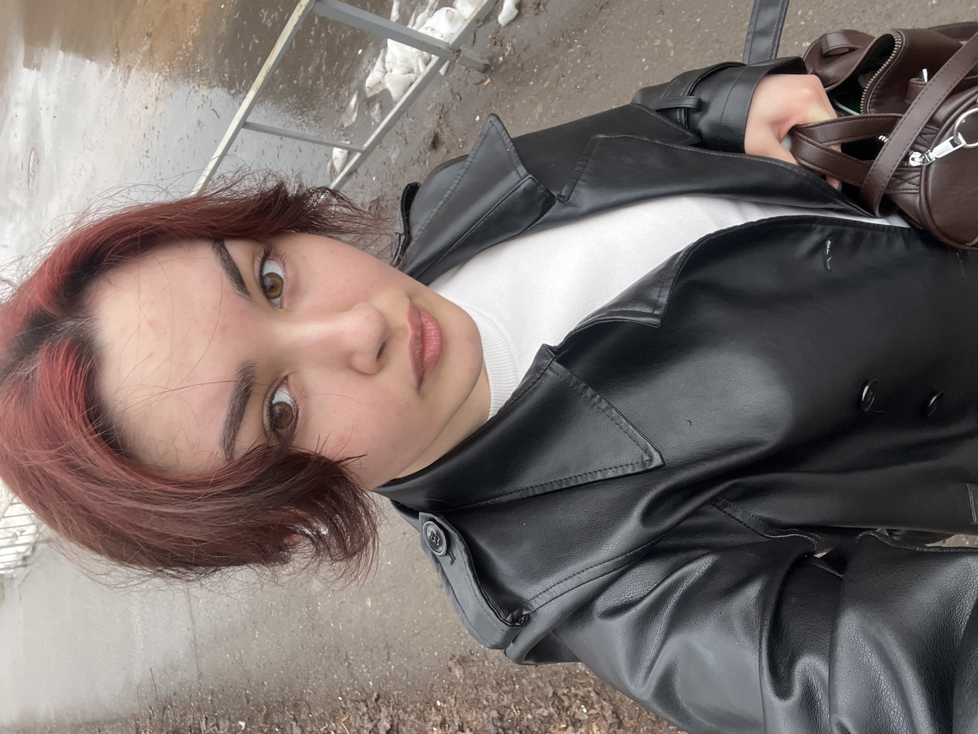 A young woman stands outdoors on a wet pavement area that suggests recent rain or melting snow. Her hair is auburn. She is wearing a white sweater beneath a black leather-like jacket and a brown bag. She is holding the brown bag. The background reveals a metal fence, patches of snow, and evidence of street debris or leaves scattered on the ground.
The location is not possible to confirm using only this image.