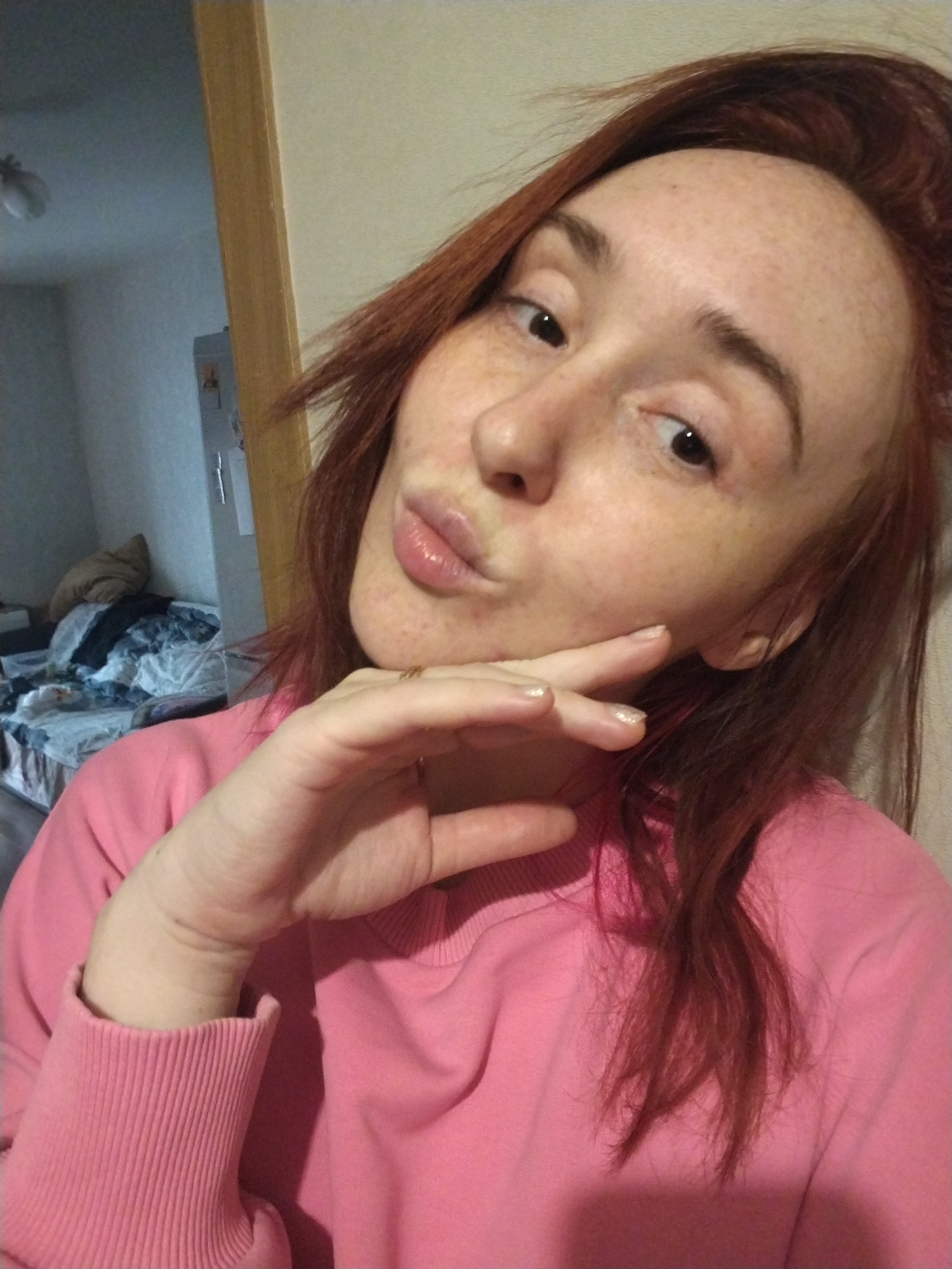A young woman with red hair and freckles is taking a selfie. She is wearing a pink sweatshirt and holding her hand to her cheek. Her lips are pursed. The background is a cluttered room with a bed and a partially visible doorway. The location appears to be an interior residential space. While the location is unknown, there is nothing in the image to suggest it is in Izhevsk, Russia.
