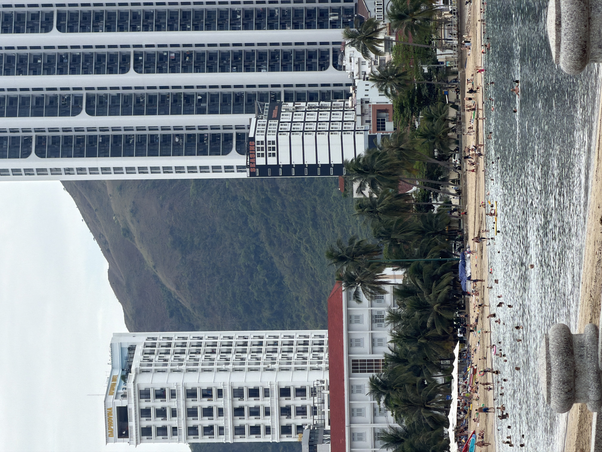 The image shows a beach scene in Nha Trang, Vietnam. The water is a reflective gray-blue and the sand is light tan. Many people are in the water and on the beach, some swimming and some lounging. A line of palm trees runs along the beach. Behind the palm trees are several tall, white buildings and a green, mountainous hill. The sky is overcast. A gray, stone barrier is partially visible on the right side of the image.
