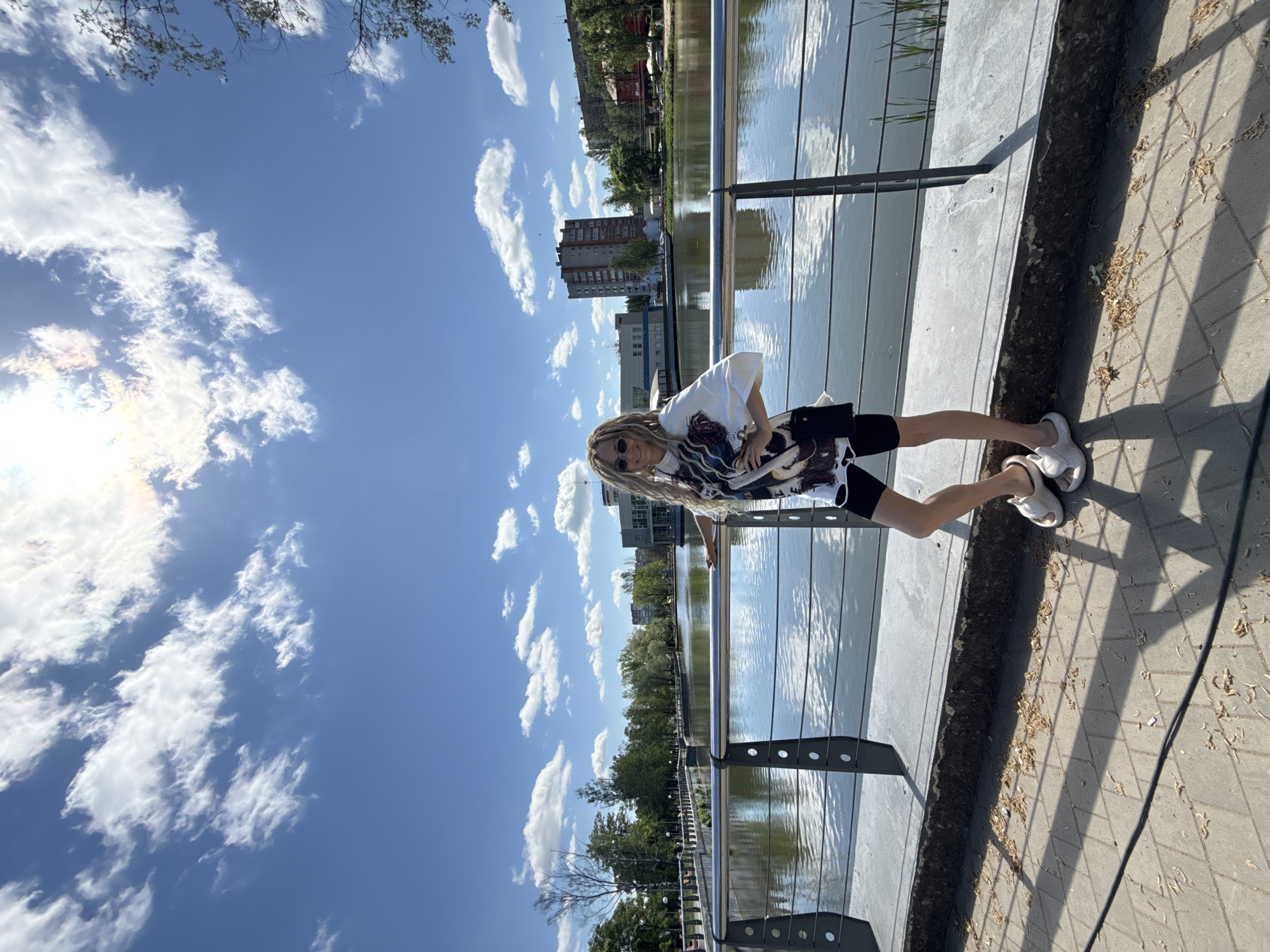A woman stands on a paved platform next to a body of water in a city identified as Unknown, Unknown. She wears a white t-shirt, dark shorts, white sandals, and sunglasses, with long hair. A metal railing separates the platform from the water. In the background, buildings, trees, and a blue sky with scattered clouds are visible.