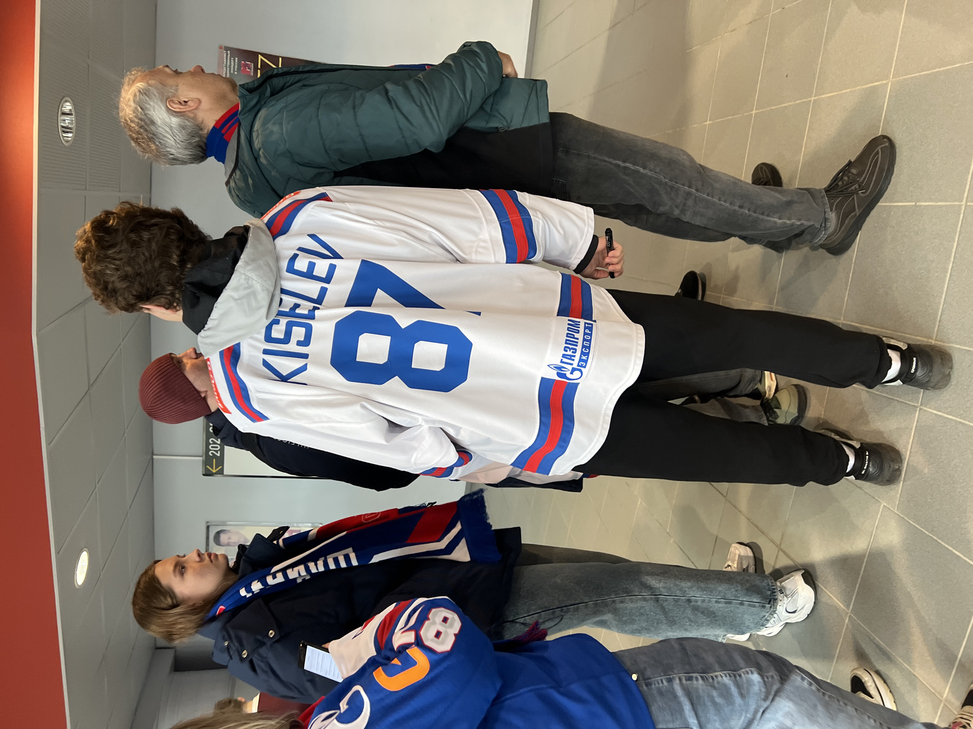 A group of people are standing in an indoor area, likely a stadium or arena concourse, with a sign indicating "202."
The people are wearing casual clothing. One person is wearing a white hockey jersey with blue and red stripes and the name "KISELEV" and the number "87" on the back. Another person is wearing a blue scarf and a blue hockey jersey with an orange "87." An older man is wearing a dark green jacket and a scarf with red, blue, and white stripes.
The location is indicated by the "Gazprom" logo on the jersey, suggesting Saint Petersburg, Russia, as Gazprom sponsors the hockey club SKA Saint Petersburg.