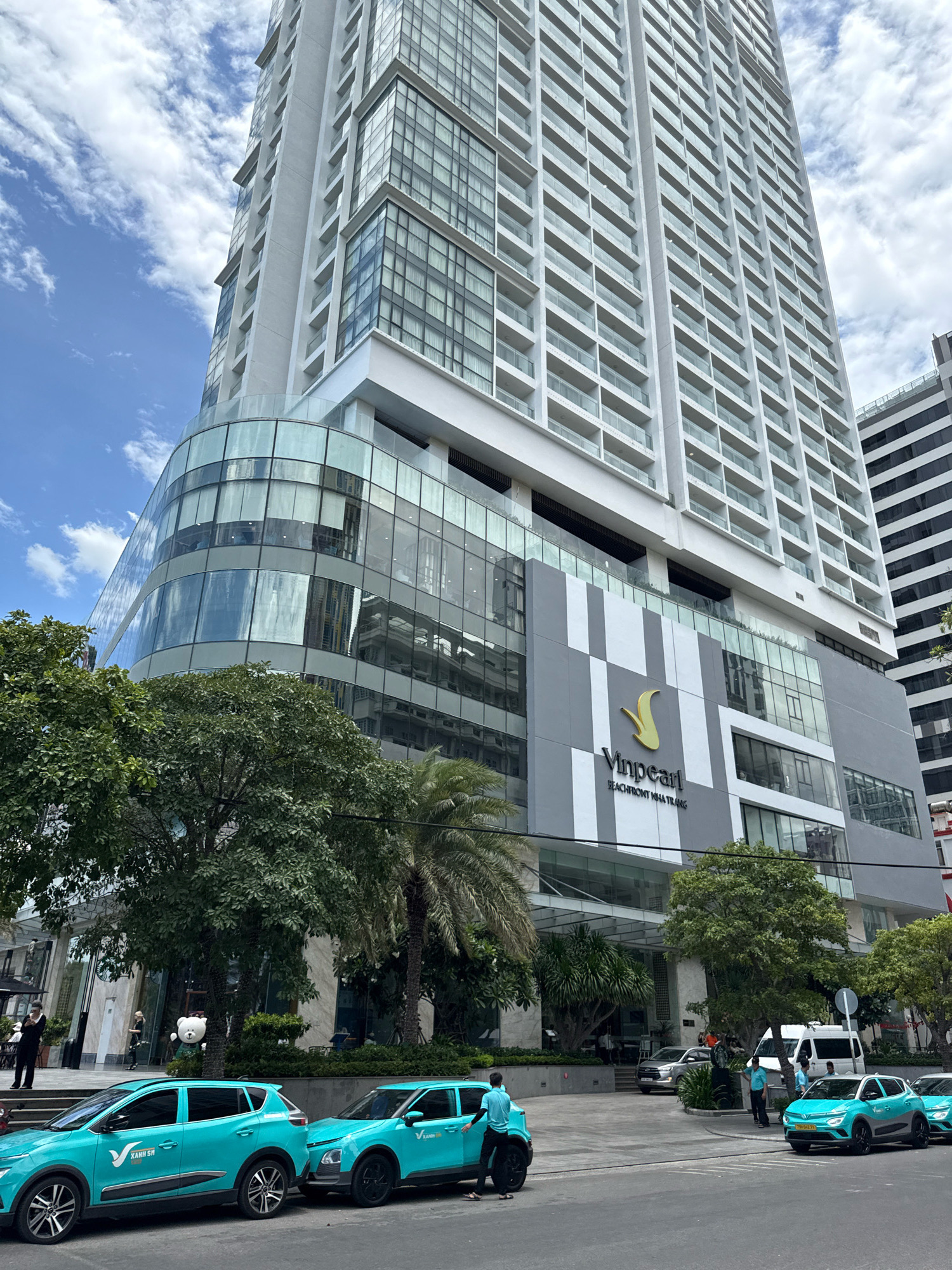 The image shows the Vinpearl Beachfront Nha Trang hotel, a tall, modern building with a curved glass facade at its base. The hotel's logo, a gold stylized 'V', is prominently displayed on a grey and white patterned section of the building. In front of the hotel, several bright teal colored vehicles, identified by the VinFast logo and the text "XANH SM" on their sides, are parked or driving on the street. These are likely electric taxis, part of a ride-sharing service. Lush green trees, including palm trees, are visible in front of the hotel, suggesting a tropical or subtropical urban environment. The sky is blue with scattered white clouds. People are seen walking on the sidewalk and near the hotel entrance, and some are interacting with the vehicles. The overall scene depicts a busy urban area with a focus on modern architecture and transportation infrastructure in Nha Trang, Vietnam.