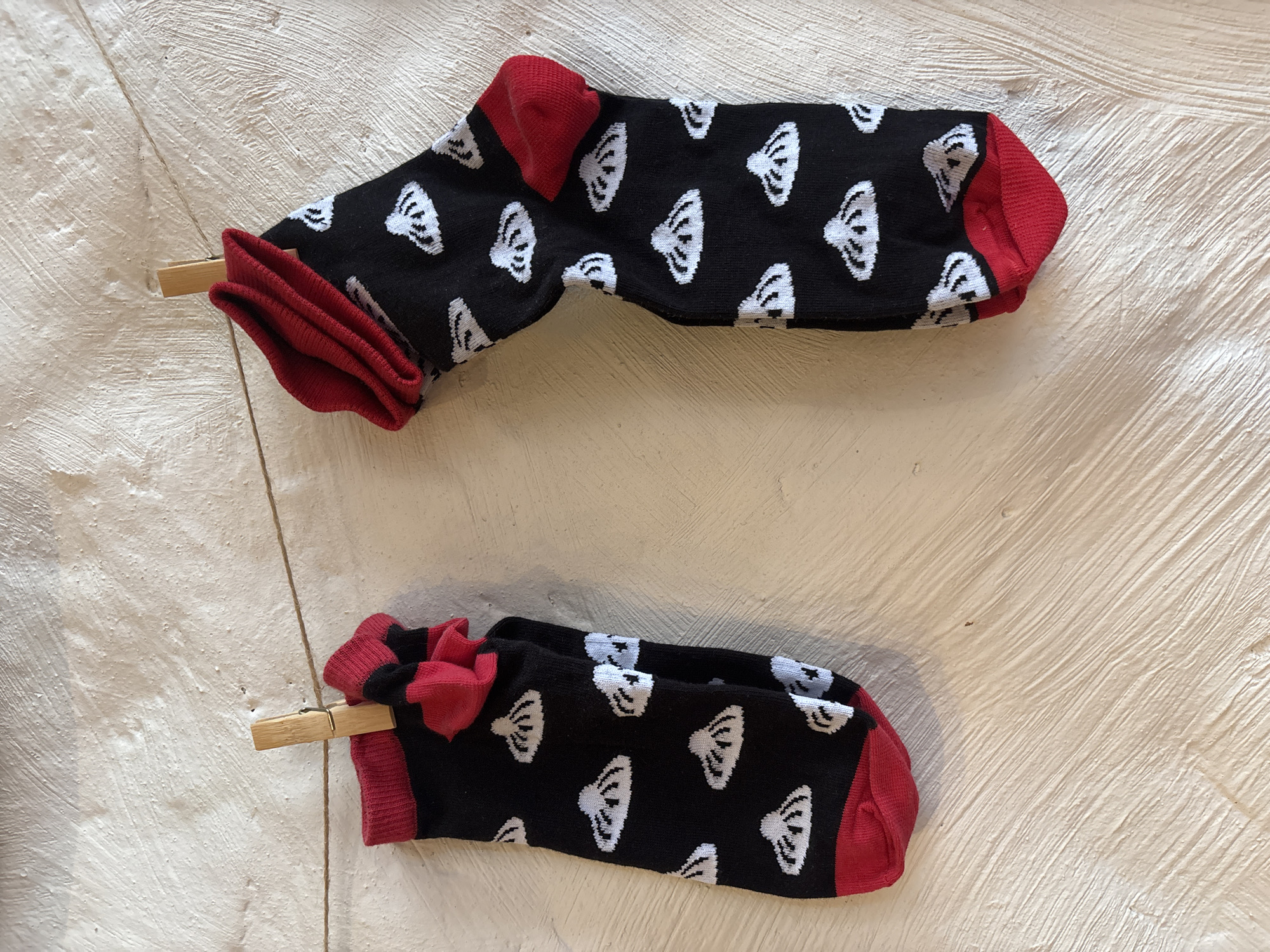 Two socks are laid out on a lightly textured white surface. Each sock is primarily black, with red accents at the heel, toe, and top cuff. The socks have a repeating pattern of stylized white shapes resembling stylized mouse ears. Each sock is clipped at the cuff with a wooden clothespin. The description does not include any information about the location in the city of Izhevsk, Russia.