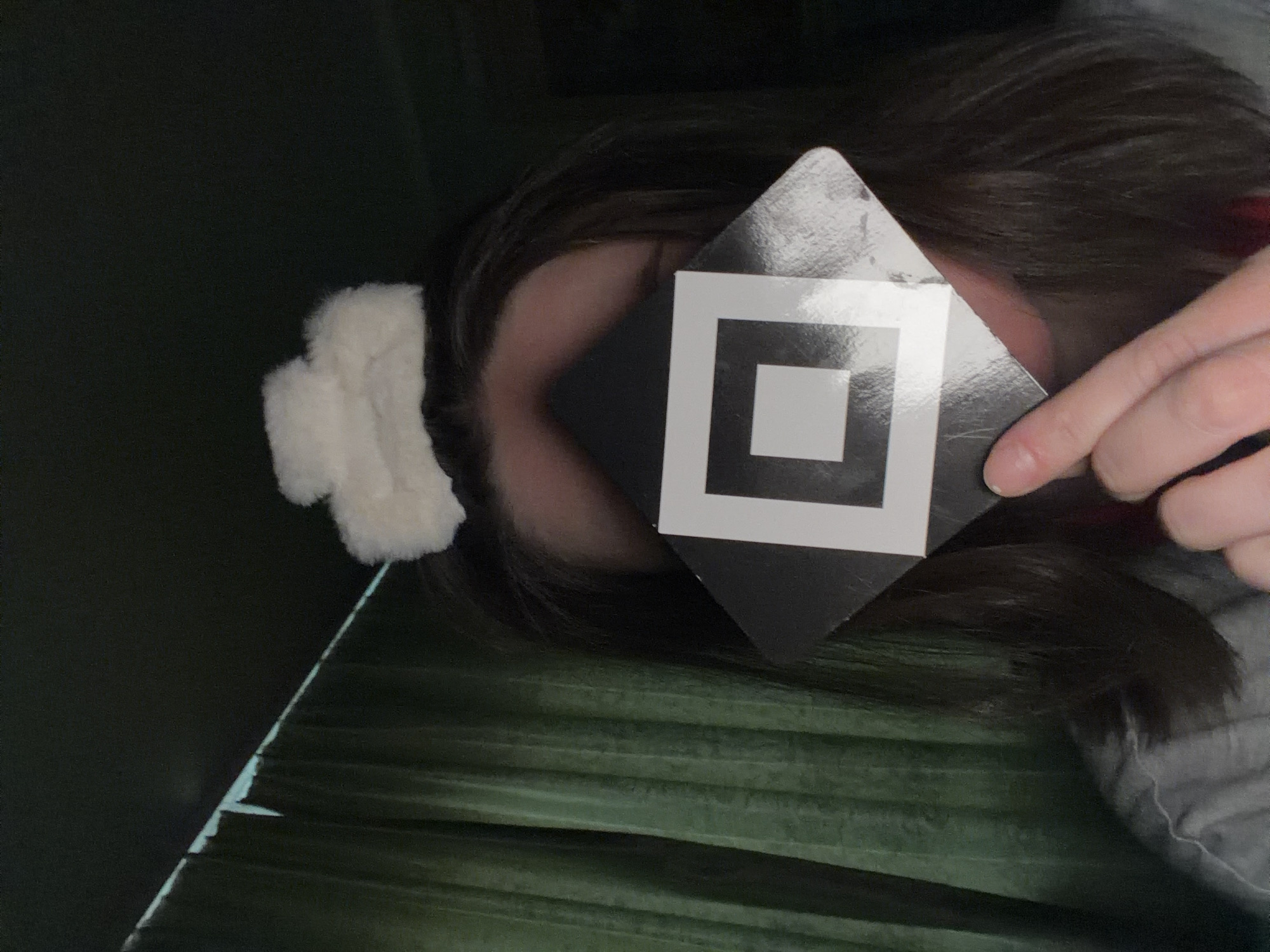 A person lies on a green, ridged surface, possibly a sleeping bag or mattress. They hold a square black card with a white square in the center over their forehead, obscuring their eyes. The person has dark hair, and a white, fluffy object is visible on the left side of their head, possibly an ear muff or decorative hair accessory. The location and city are unknown.