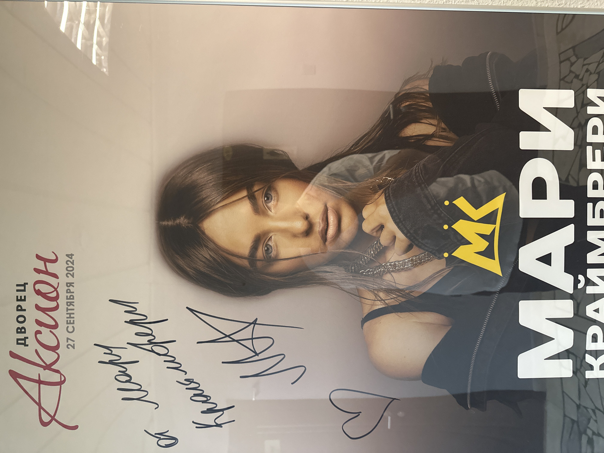 The image shows a poster of the singer Marie Kraimbrery. The poster includes the name of the venue "Дворец Аксион" and the date of the concert "27 сентября 2024" (September 27, 2024). Several signatures are visible in the bottom left of the poster. The location is Izhevsk, Russia, as indicated by the name of the venue.