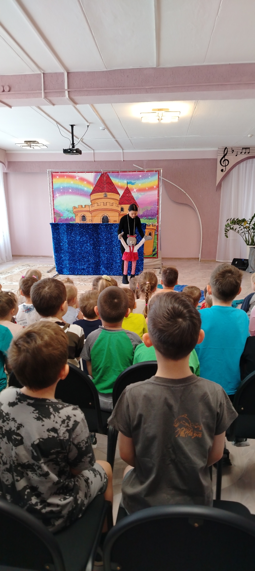 Captured in Chaikovsky, Russia, an audience of children is seated indoors, attentively watching a puppet show. The show is presented on a stage featuring a castle backdrop and a blue curtain. A person dressed in black is operating the puppet, which is dressed in a red outfit. The room's decor includes a light pink wall paint, a musical note decoration, and a potted plant. A projector is mounted on the ceiling.