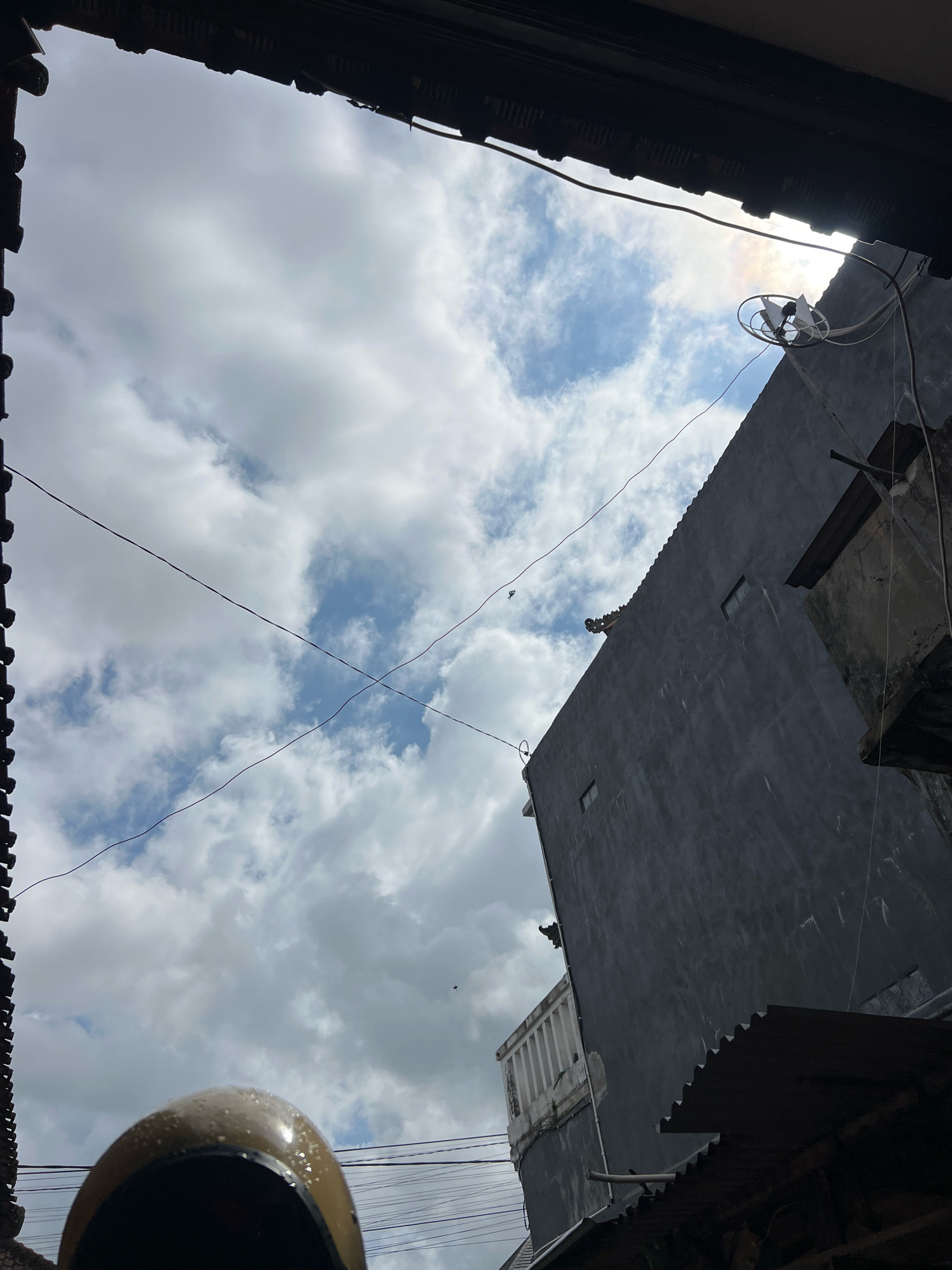 A low-angle, upward-facing shot captures a cloudy sky above urban architecture in Denpasar, Indonesia. The foreground is partially obscured by the dark, textured eaves of a roof, from which a portion of a light brown helmet, possibly with water droplets, is visible at the bottom left. Diagonal black electrical wires traverse the sky. A tall, dark gray building with a corrugated metal roof dominates the right side of the frame. The building has at least two small, dark rectangular windows. A white satellite dish with two propellers is mounted on the building near the top right, with the sun partially visible behind it, creating a slight lens flare effect. At the peak of the building's roof, a decorative, possibly stone, gargoyle-like figure is visible. Another building, lighter in color with a white railing, is situated to the left of the gray building. The overall scene suggests an overcast day in a densely built urban environment.