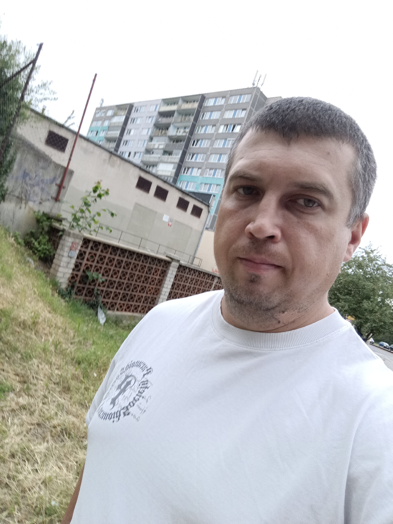 The image is a selfie taken outdoors with a man in the foreground wearing a white t-shirt. Behind him, a multi-story apartment building with numerous windows is visible, along with a smaller, lighter-colored building and a decorative brick wall with a patterned design. The ground is covered in grass and dry vegetation. A fence post and some greenery are also present in the background. The scene appears to be in a residential area.