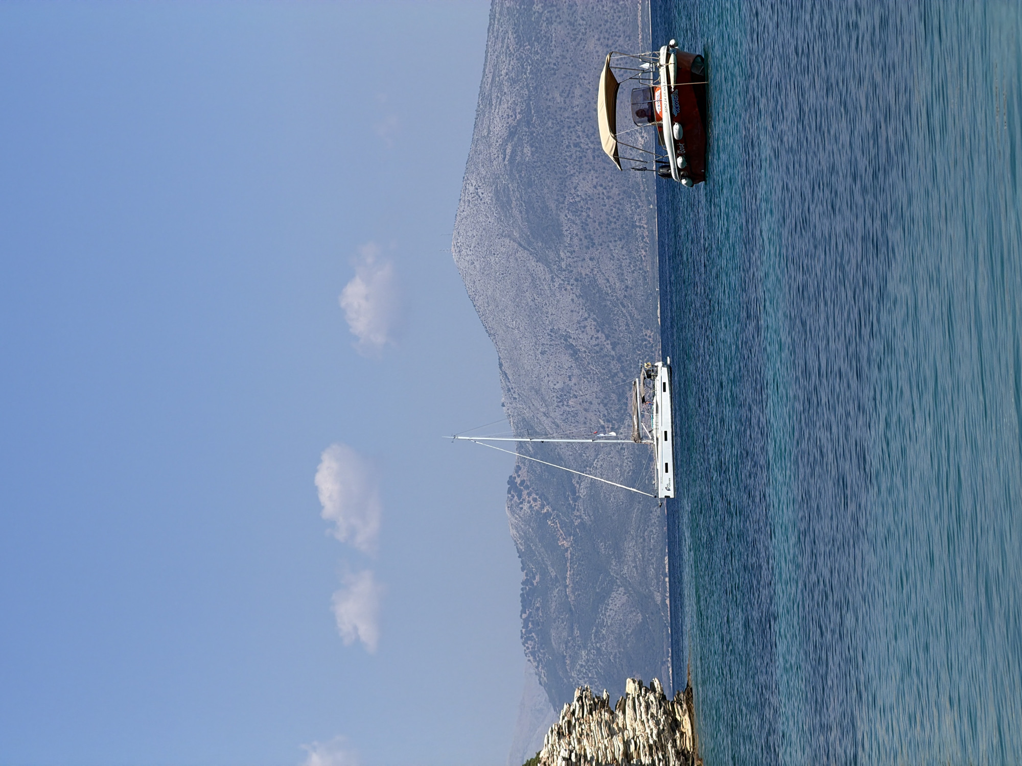 The image depicts a serene coastal scene in the Municipal Unit of Kastos, Greece, on a clear day. The foreground is dominated by tranquil, azure-blue waters, with gentle ripples reflecting the sunlight. In the middle ground, two boats are anchored. On the left, a white sailboat with a tall mast stands gracefully, its hull visible just above the waterline. To the right, a smaller red motorboat with a beige canopy is positioned, featuring a person at the helm. The background is characterized by rugged, sparsely vegetated mountains, their slopes textured with small bushes and rocks, under a bright blue sky dotted with a few wispy white clouds. A distinctive rocky outcrop, characterized by its layered strata, juts out from the left side of the frame, adding a dramatic geological element to the landscape.