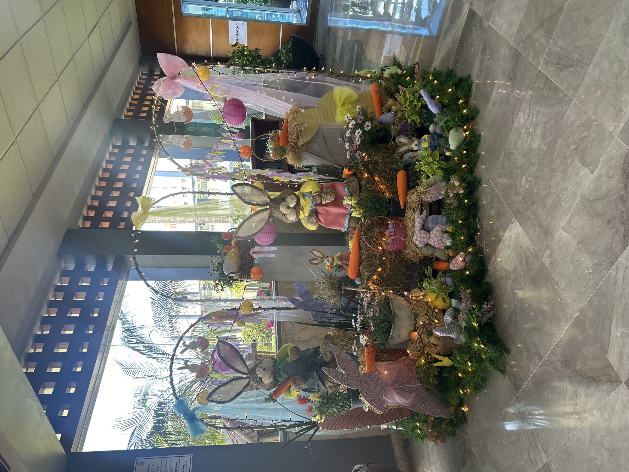 The image shows an Easter-themed decoration in what appears to be a lobby or entrance area. The decoration features multiple stuffed bunnies of various sizes and styles. There are also artificial carrots, flowers, and green foliage arranged around the bunnies. The display is surrounded by small string lights that are illuminated. The decoration is placed on a tiled floor. The lighting is bright and the overall impression is festive. While I can infer the decorations are within an indoor space, I have no information to support that the decorations are within the city of Puebla, Mexico. I can only describe what is visible in the image.