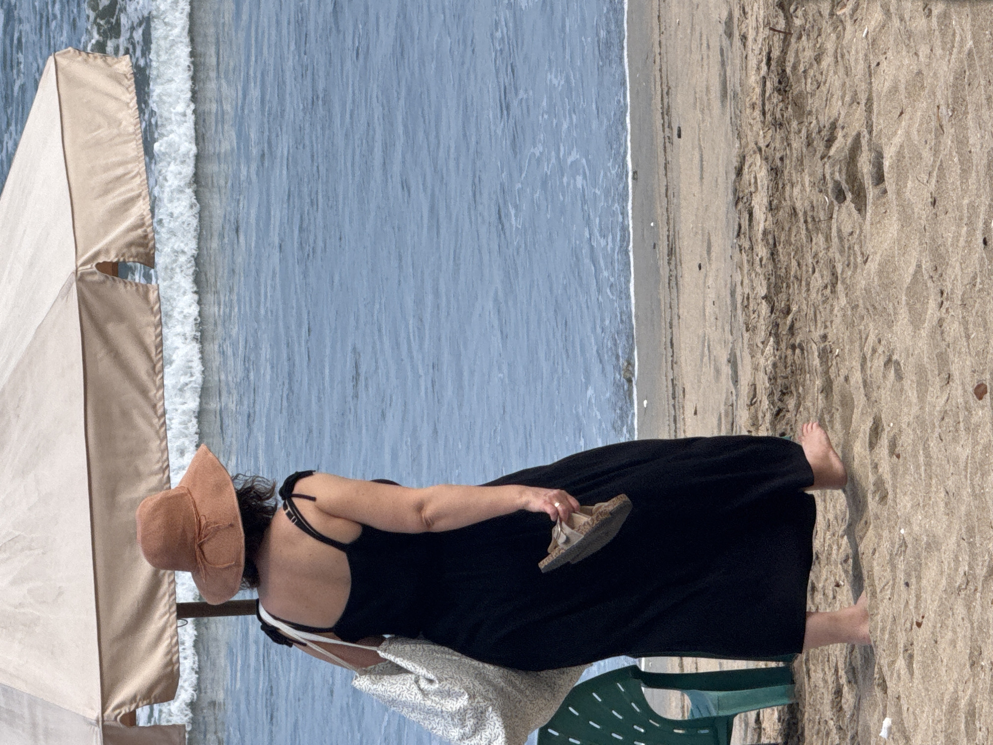 A person wearing a wide-brimmed, tan hat and a long, black dress is walking on a sandy beach in Kuta, Indonesia. The person is carrying a white tote bag with a floral pattern on their left shoulder and a brown sandal in their right hand. They are barefoot, and the sand shows footprints. In the background, the ocean has moderate waves, and a tan beach umbrella is partially visible. A green plastic chair is to the left of the person.