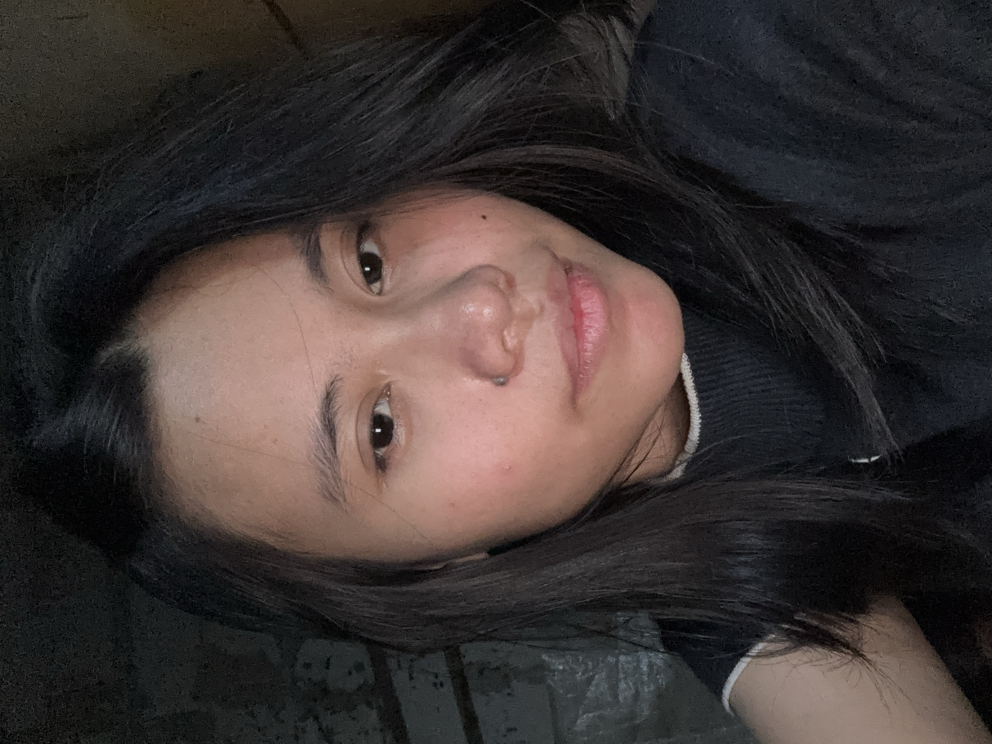 The image shows a close-up, frontal view of a young East Asian woman's face. She has dark hair that falls past her shoulders and is wearing a dark-colored top with a white trim. Her skin appears smooth with some subtle blemishes. She has a small piercing on her left nostril and a beauty mark on her right cheek. Her eyes are dark brown and she has a neutral expression. The background is dark and out of focus, with some textures visible that might suggest a wall or a distressed surface. The lighting is dim, casting shadows on her face and creating a slightly grainy effect. The location is stated to be East Jakarta, Indonesia, though no specific landmarks or environmental cues within the image directly confirm this.