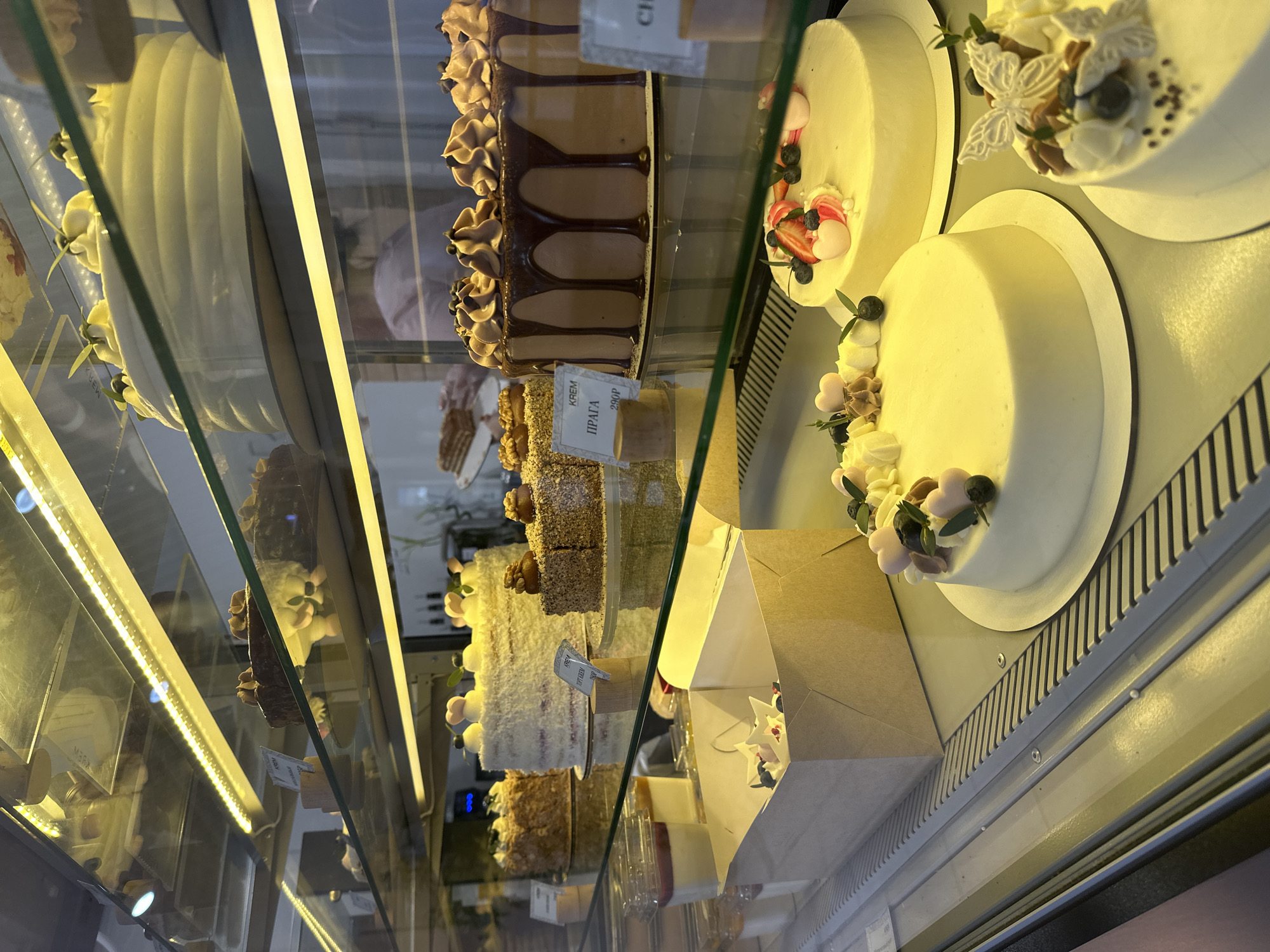 The image displays a brightly lit glass display case inside a confectionery or bakery in Votkinsk, Russia, showcasing multiple tiers of cakes and individual desserts.
On the top shelf, several large, elaborately decorated cakes are partially visible, one featuring white frosting with green foliage elements, and another with layered white frosting.
The middle shelf presents a variety of cakes: a large, round cake with textured white frosting and dark berries; a square cake topped with ground nuts; and a prominent round chocolate cake with dark chocolate ganache dripping down its sides and piped chocolate rosettes on top. A small, wooden stand next to a different cake holds a white price tag labeled "КРЕМ ПРАГА" (KREM PRAGA) and "290P," indicating "Prague Cream" and a price of 290 Rubles. Additionally, clear plastic cups contain individual layered desserts, and an open, light brown cardboard box containing white, star-shaped decorations is present.
The bottom shelf features three large, round cakes with smooth, light-colored frosting. One cake is adorned with fresh strawberries, blueberries, and small pink heart-shaped candies. Another is decorated with small, light pink flower-like elements and blueberries. The third cake includes white butterfly decorations and dark sprinkles.
The interior of the display case is illuminated by horizontal LED strip lighting embedded in the shelves. A blurred reflection of an individual wearing a light-colored, possibly pink or purple, top is visible in the background glass. The base of the display case is a ribbed, metallic surface. No specific actions or interactions are depicted.