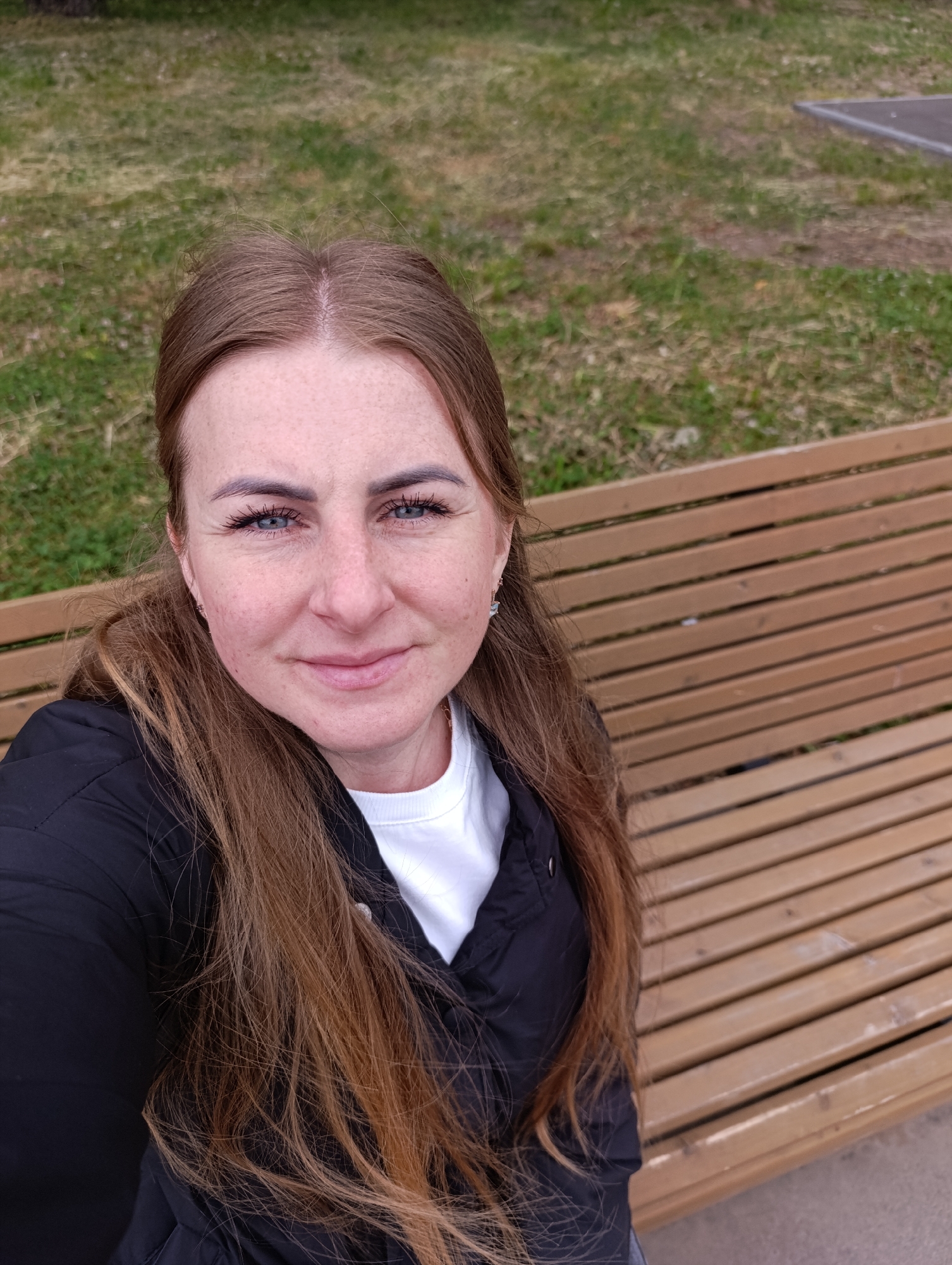 A woman is seated on a brown wooden park bench. She has light brown hair with darker roots, blue eyes, and freckles. She is wearing a black jacket over a white top. Behind her is a grassy area with patches of dry grass and some concrete. The location is in a city named Unknown, Unknown.