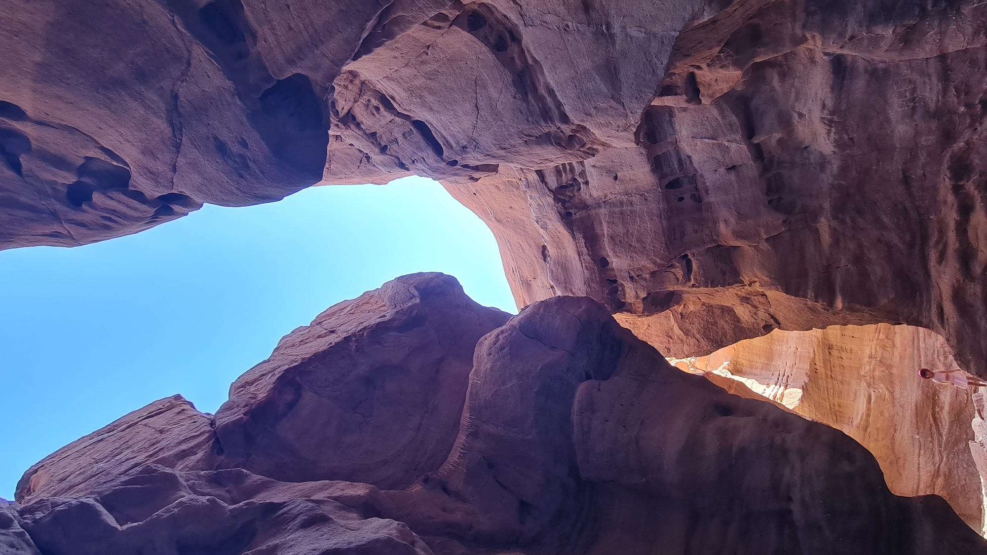 A low-angle shot shows a sandstone canyon in the city of Hevel Eilot Regional Council, Israel. A wide opening in the canyon roof allows a clear blue sky to be seen. The rock formations are textured with holes and crevices. A person is standing at the top of a rock formation on the right side of the frame, near an opening that lets sunlight through.
