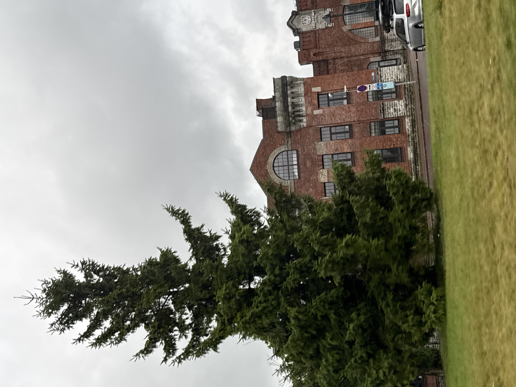 A tall, dark green evergreen tree stands in the foreground on the left side of the image. Behind the tree, to the right, is a red brick building with arched windows and some stone accents. The sky is overcast and grey. In front of the building, a silver station wagon with a roof rack is parked. The ground in front of the tree is covered in green grass, and a paved area is visible between the grass and the building. The scene appears to be in an urban setting, with the building likely being a historical structure given its architecture. This building is identified as the "Bunkker" Hotel, located in Tallinn, Estonia.