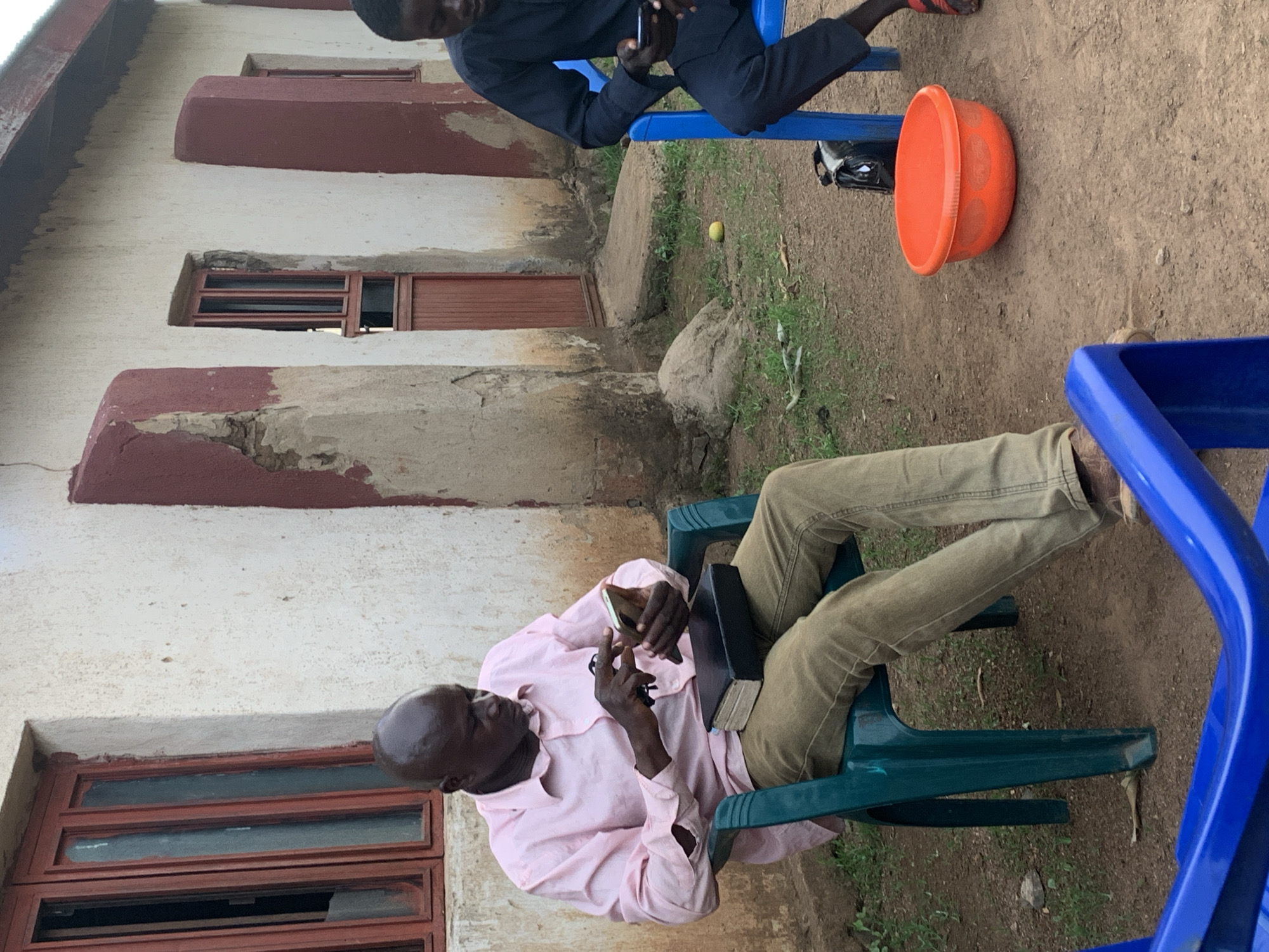 Two African men are seated on blue and green plastic chairs outside a building. One man, wearing a pink shirt and khaki pants, sits on the green chair holding a smartphone and a book. The other man, wearing a black top and pants, sits on the blue chair looking at his smartphone. An orange bowl is on the ground near them. The building is white with red trim around the windows. There is dirt ground and some greenery around the building. An identification of the city of Agban is not possible based solely on the given image.