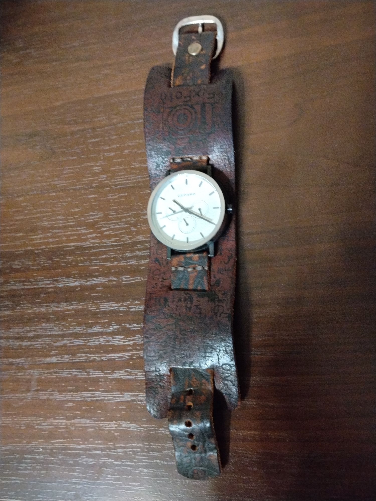 A vintage, brown leather cuff watch with a white face and silver hands is displayed on a dark brown, horizontally-grained wooden surface. The watch band features embossed text and a buckle closure. The background is a plain wooden table. There is no indication of the city of Krasnogorsk, Russia, or any people present in the image.