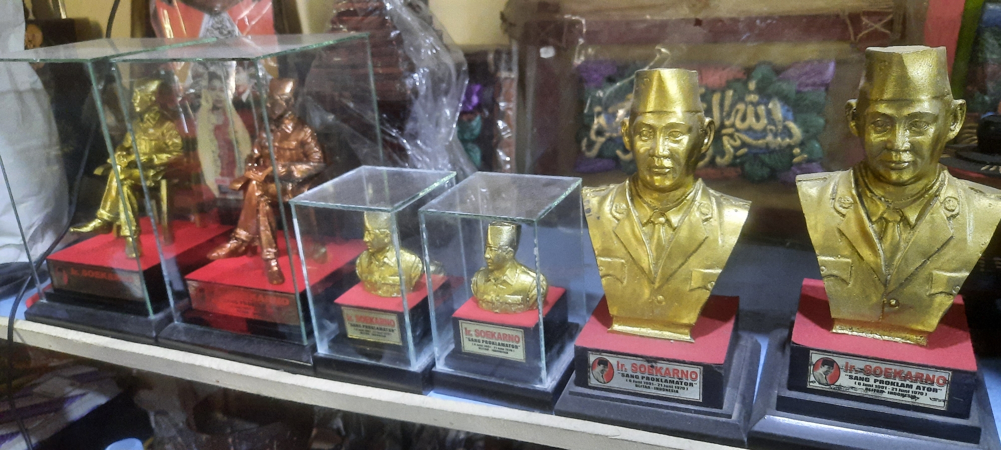 In Blitar, Indonesia, a shelf displays several gold-colored busts and figurines of Ir. Soekarno, the first president of Indonesia. The items are placed in glass cases and on individual black bases with red platforms. Some of the busts have small plaques with text that appears to be biographical information about Soekarno. In the background, there are various other decorative items and some plastic-wrapped objects.