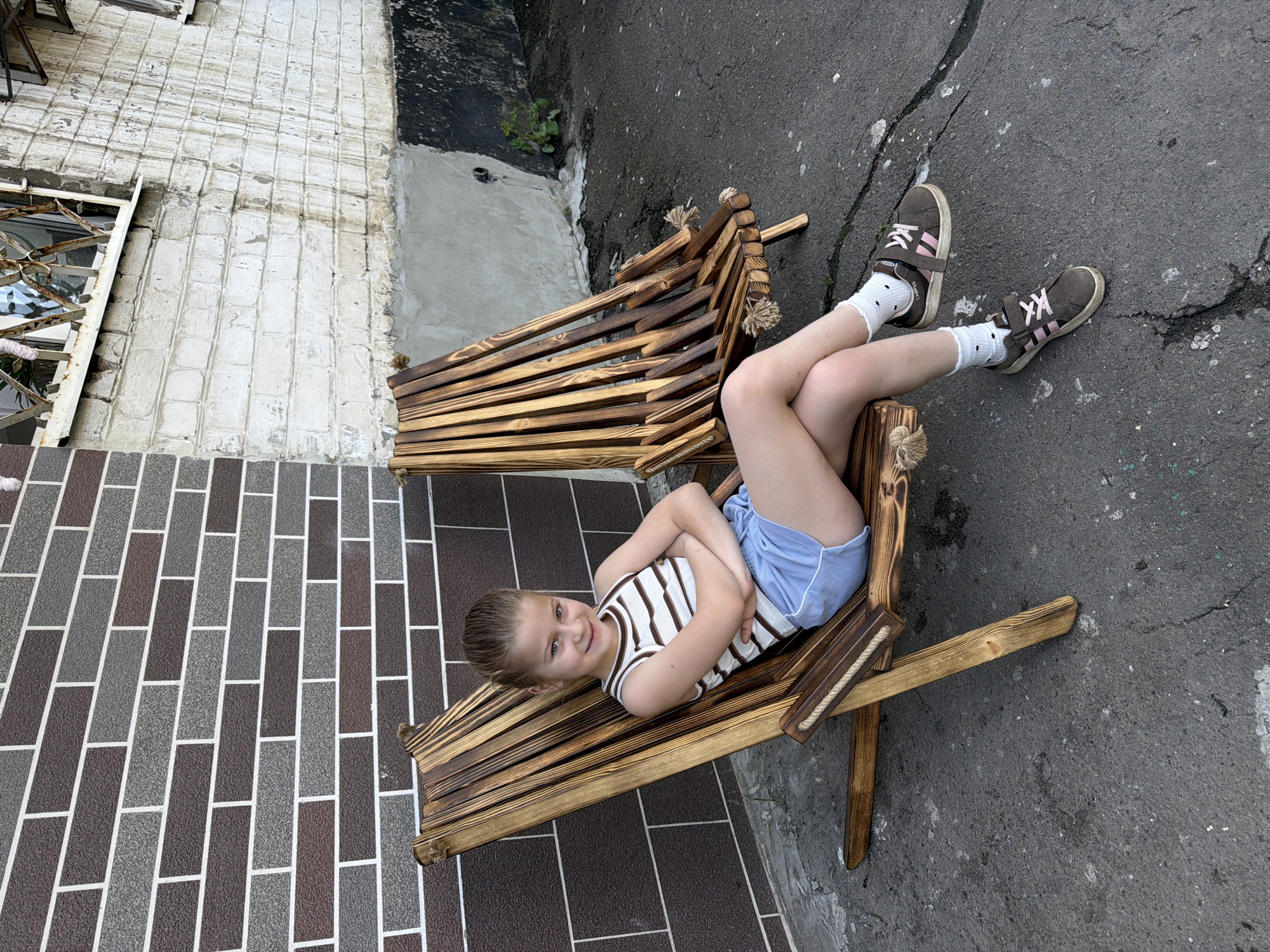 A young girl with blonde hair pulled back is sitting in a wooden lawn chair outdoors. She is wearing a white and brown striped tank top and light blue shorts. Her arms are crossed and she is looking at the camera with a slight smile. Her legs are crossed at the ankles. She is wearing white socks with black polka dots and brown sneakers with pink laces. Behind her, another identical wooden lawn chair is visible, empty. The background consists of a dark brown brick wall on the left and a light grey, weathered brick wall on the right. The ground is asphalt with some cracks and debris. There is a small green plant growing near the base of the grey wall. The scene appears to be in an urban setting.