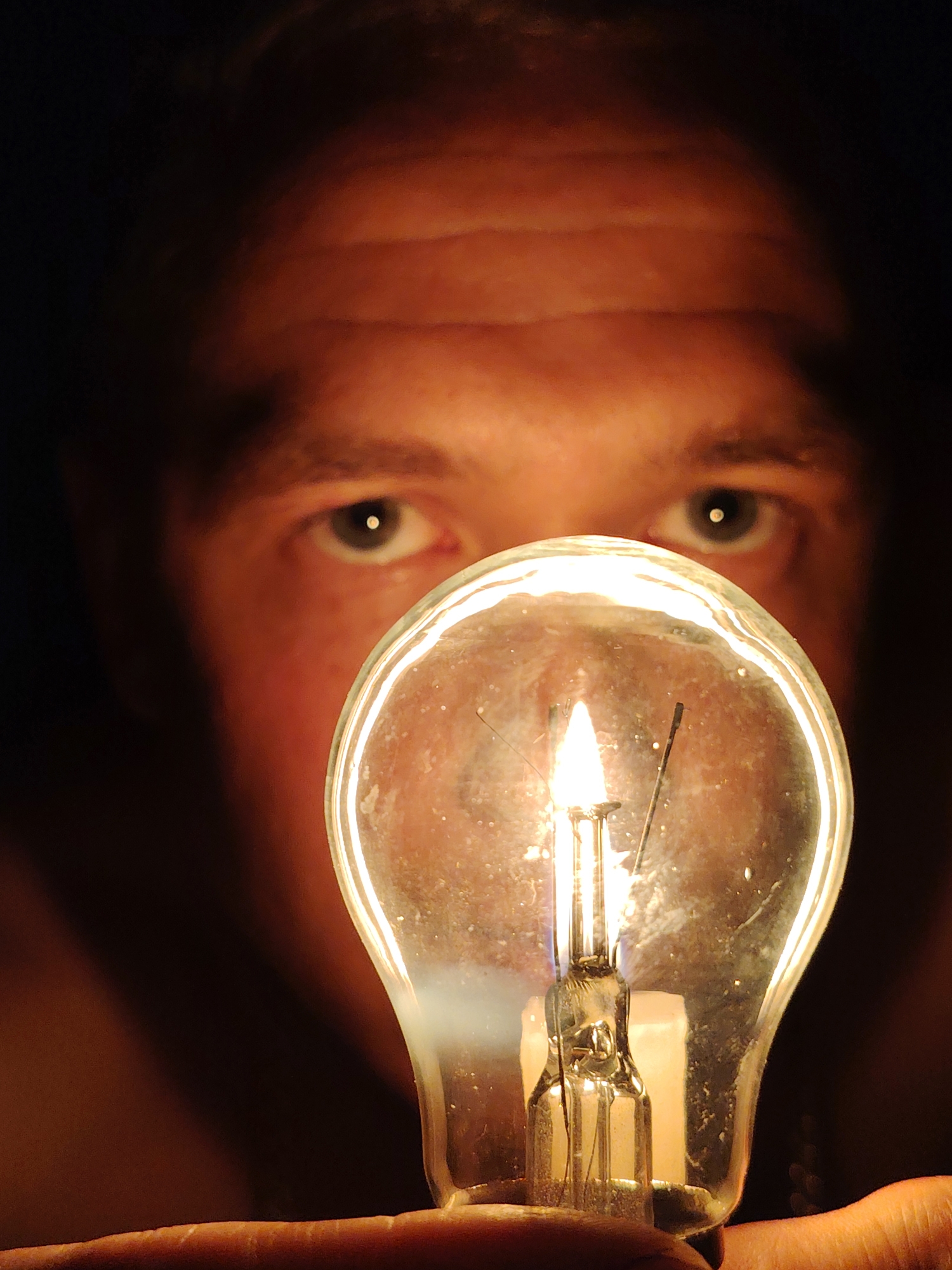 A close-up, low-angle shot shows a person's face in the background, out of focus, with their eyes visible. In the foreground, a lit incandescent light bulb is held by fingers, with the filament glowing brightly. The background is dark, suggesting a dimly lit environment. The image is centered on the light bulb, with the person's face serving as a backdrop. The lighting is warm and intense due to the bulb. No specific location in Chaikovsky, Russia can be determined from the image.