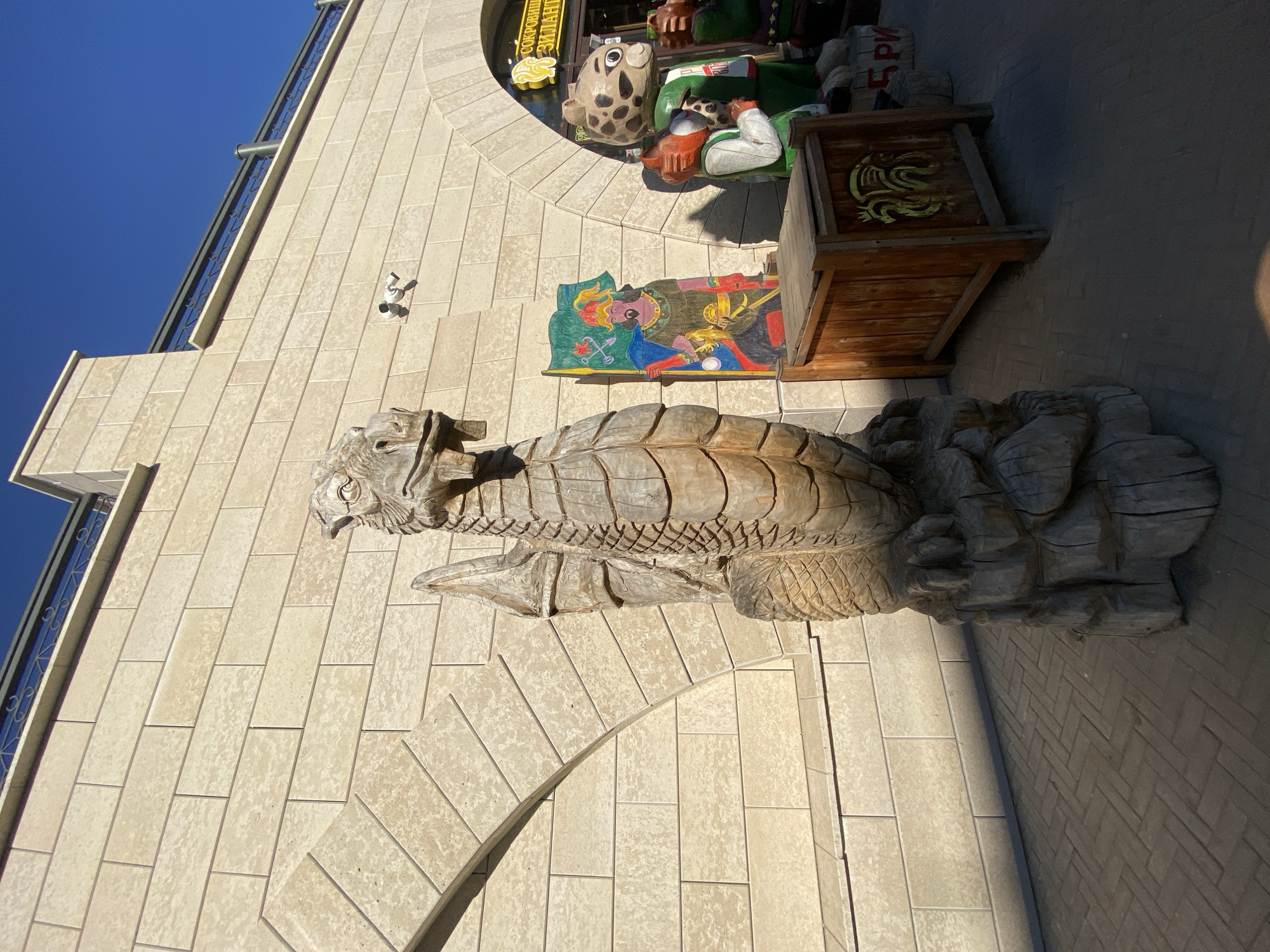 The image shows a building facade in Kazan, Russia. A large wooden dragon sculpture is affixed to the wall. To the right of the dragon, are various wooden figures and signs. The sky is blue and the building is constructed from light-colored bricks.