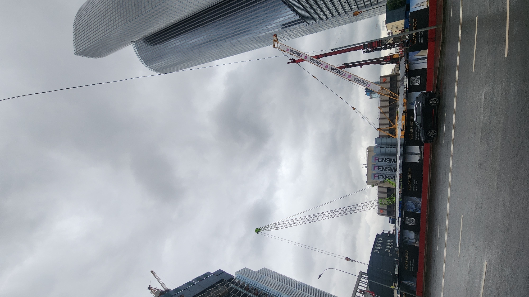 The image captures a construction site in Moscow, Russia, under a cloudy sky. The foreground shows a wet asphalt road with white lane markings. A black SUV is parked on the right side of the road, behind a temporary construction barrier. The barrier features advertisements for "SEZAR GROUP" and images of buildings.
Behind the barrier, the construction site is dominated by several large cranes. One tall, slender crane with a green top extends upwards, while another, a larger, more robust crane, is red and white with the text "FENSMA" visible on its boom. The site itself is a hive of activity, with unfinished structures and scaffolding evident. In the background, two modern skyscrapers with reflective glass facades rise above the construction, indicating an urban setting. The overall impression is one of ongoing development and construction within a metropolitan area.