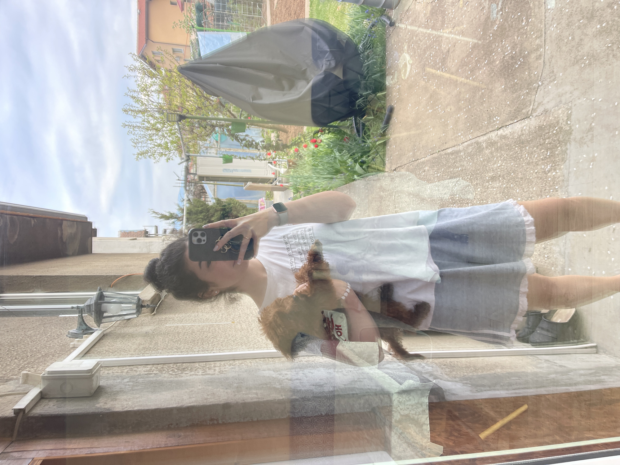 The image is a reflection in a window, showing a young woman holding a small, brown dog. The woman has her dark hair up in a bun and is wearing a white t-shirt, a grey dress with white trim, and a smartwatch. She is holding her phone, likely taking the picture. The dog has a white and red object attached to its collar. Through the window, a backyard is visible, featuring a dark grey outdoor furniture cover, red flowers, green plants, a concrete patio, and parts of a house under construction with scaffolding. A building with a red tile roof is also visible in the background. The presence of the characteristic building style suggests the photo was taken in Sofia, Bulgaria. Also the writing on the object attached to the dog could be in Bulgarian or another Cyrillic alphabet. In the bottom left of the reflection is a light fixture attached to a wall.