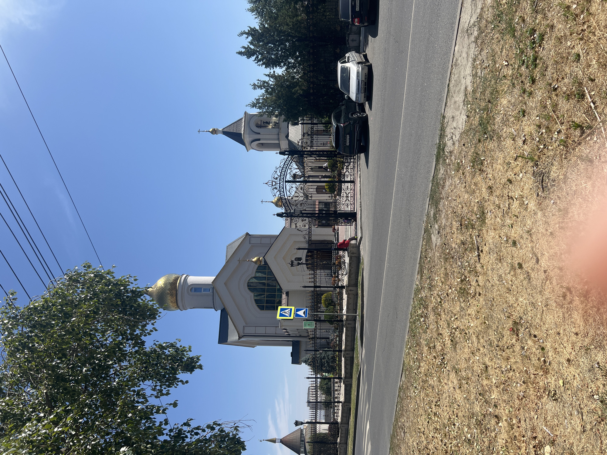 The image depicts the Church of the Archangel Michael in Volgograd, Russia, located on the Mamayev Kurgan memorial complex. The church is a modern structure with a white facade, a prominent golden dome, and a bell tower with a black roof. It is surrounded by a black ornate metal fence and a paved area with several parked cars.
In the foreground, a dry, grassy area with fallen leaves is visible, bordering a gray asphalt road. A pedestrian crossing sign is placed near the entrance to the church grounds. The sky is clear and blue, suggesting a sunny day. A large tree with green leaves partially obstructs the view of the sky on the left side of the image.
The image captures a section of the memorial complex, showcasing the architectural style of the church and its surroundings. The presence of cars and a pedestrian crossing indicates the accessibility of the site to visitors.