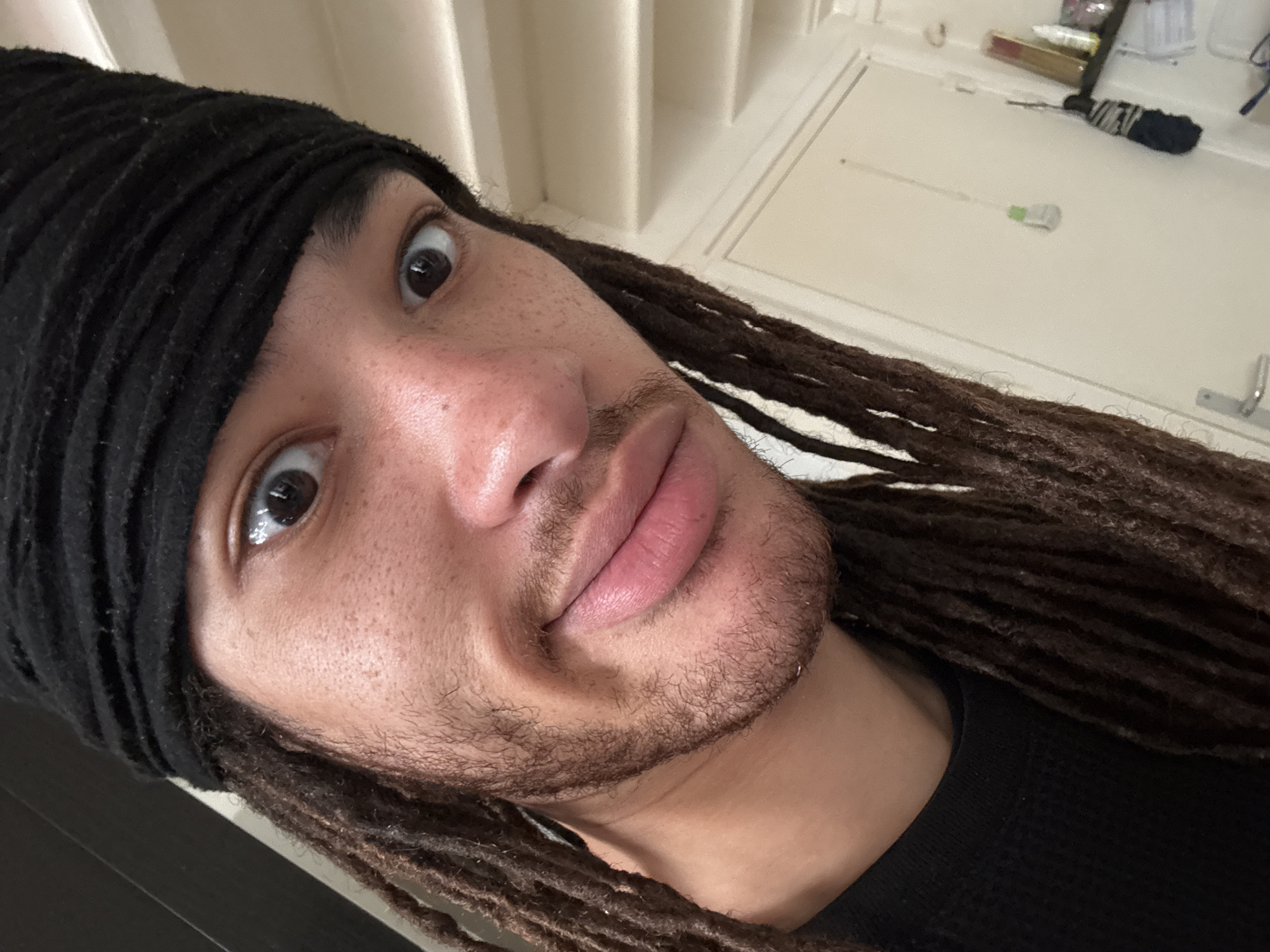 A close-up shot of a person's face, angled upwards. The person has brown eyes, light brown skin, and a short beard. They are wearing a black headband and have brown dreadlocks. The background is a white wall with vertical seams, possibly a door, and some small objects are visible on top of it. There is no information to suggest the photo was taken in Paris, France.