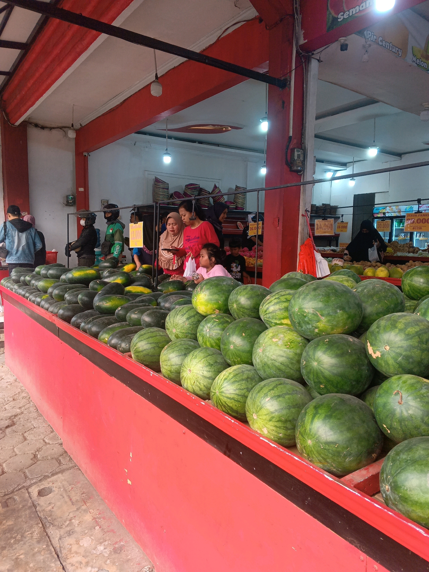 The image depicts a fruit stall in Garut, Indonesia, likely a local market or a small grocery store. The foreground is dominated by a large display of watermelons on a bright red counter. The watermelons are arranged in neat rows, with some larger ones stacked on top. Behind the counter, several people are visible. On the left, a person in a denim jacket stands looking at the produce. To the right of the counter, three women are interacting. One woman in a pink shirt is holding a phone, while another woman in a headscarf is also looking at her phone. A young girl in a pink shirt is seated nearby. Further in the background, more people can be seen, including someone in a green uniform, possibly a delivery person. The stall appears to be under a covered structure with a red and white ceiling and exposed beams. Various signs with prices and product names are visible in the background, suggesting a commercial setting. The ground is paved with what appears to be cobblestones or patterned tiles.